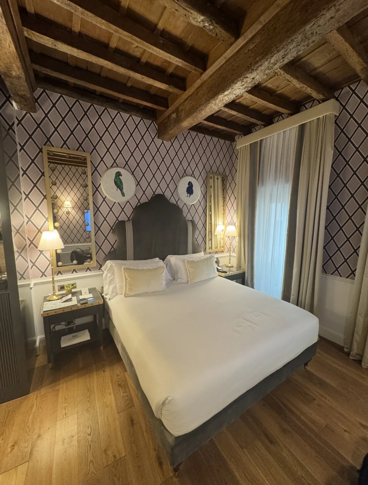 A cozy hotel bedroom with a wooden beamed ceiling, patterned wallpaper featuring diamond shapes, a large bed with white linens and pillows, two bedside lamps, a window with cream curtains, and a mirror with warm lighting on the wall.