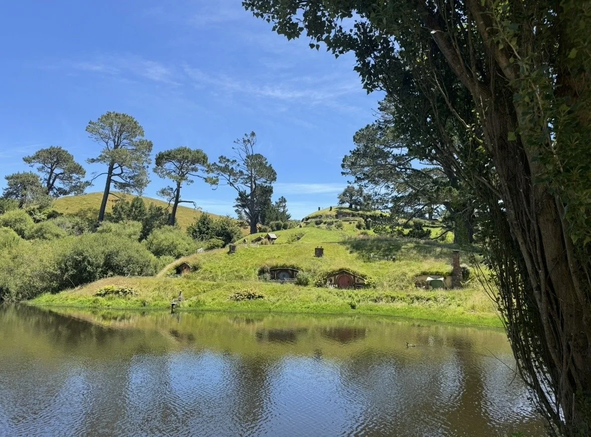 Scenic landscape with a reflecting pond, green hills, tall pine trees, and Hobbit-style houses on the hillside.