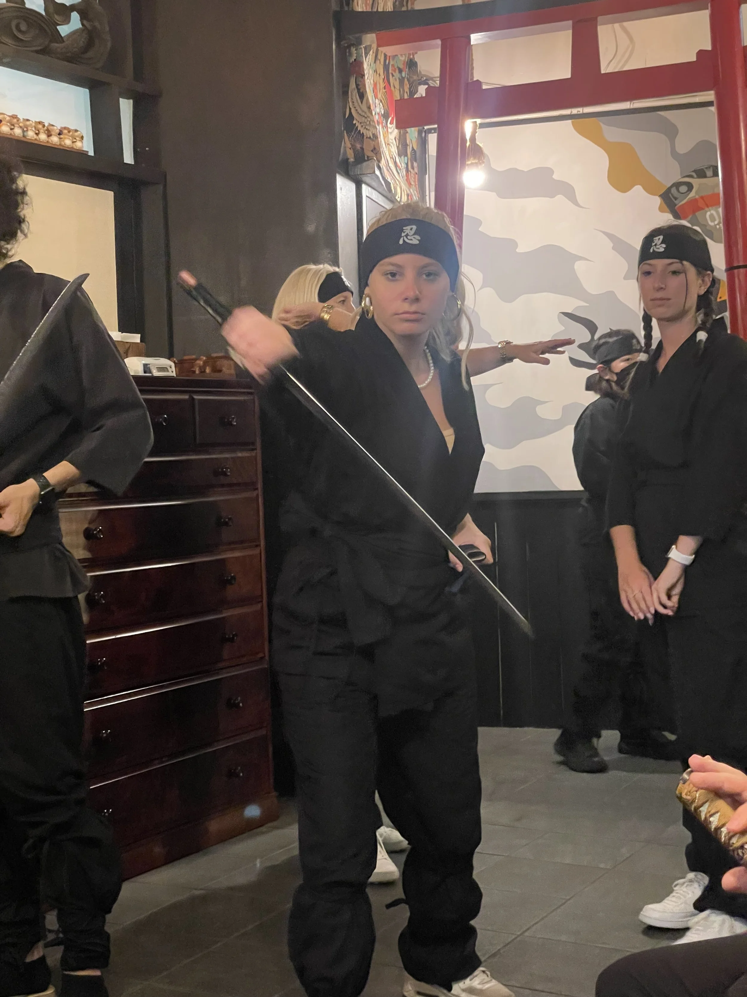 A group of people dressed as ninjas in black clothing and headbands with a ninja symbol, standing indoors with a mural on the wall and red structural elements.