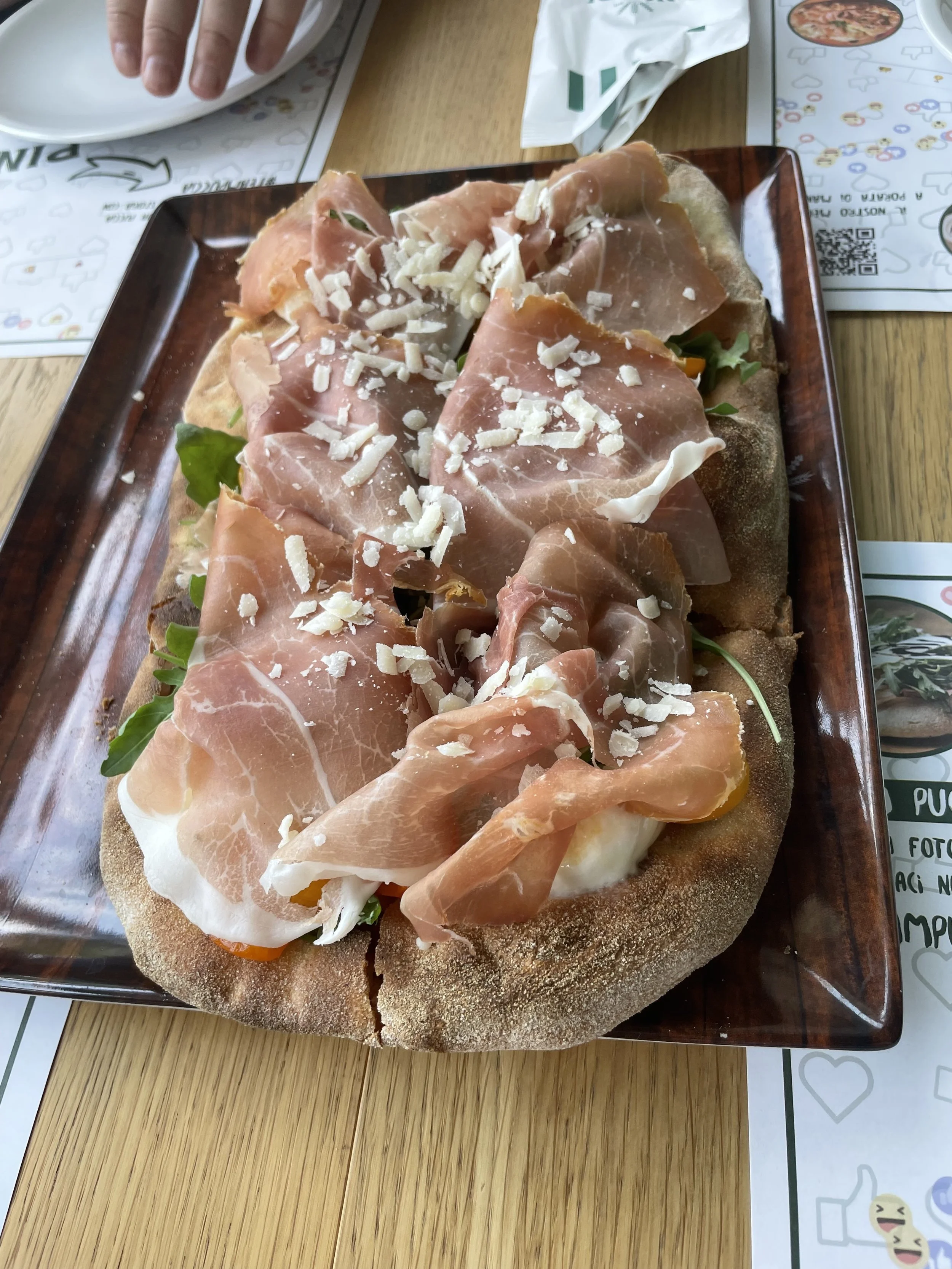 A large round pizza with a crispy crust topped with slices of prosciutto, shredded cheese, and greens, served on a rectangular wooden tray.