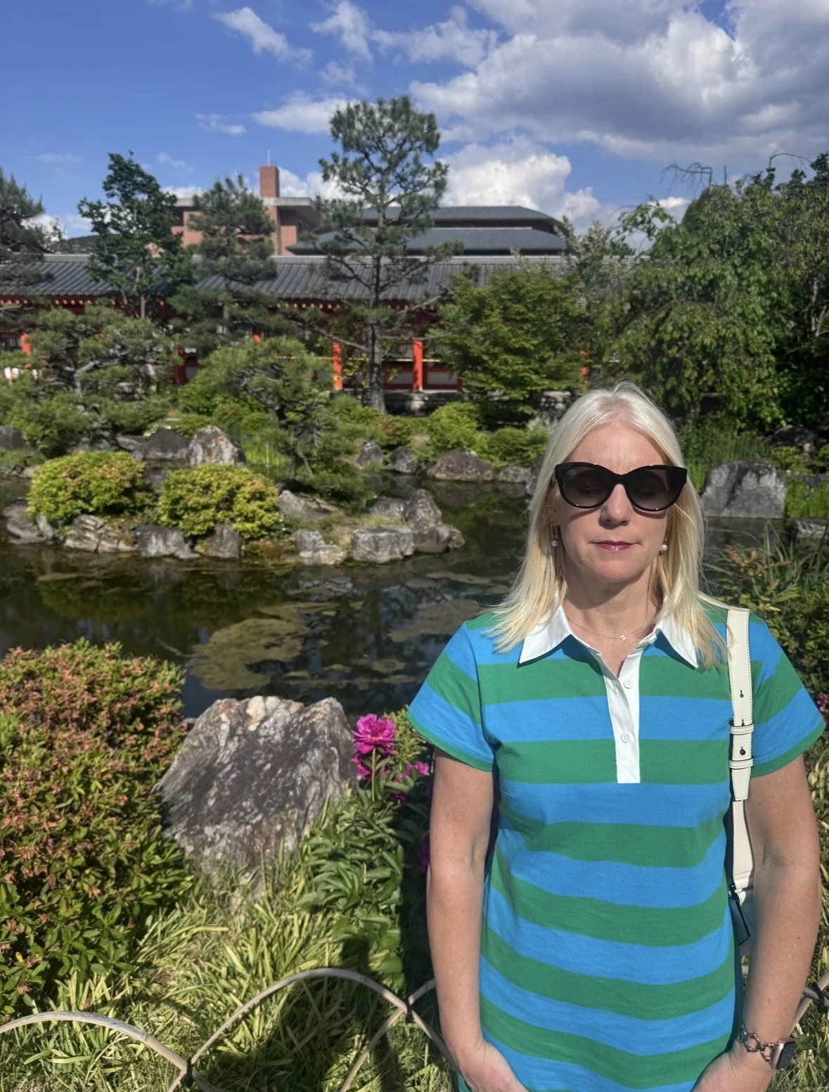A woman with blonde hair wearing sunglasses and a striped blue and green shirt, standing outdoors in a garden with rocks, shrubs, and pink flowers, with a pond and traditional Asian-style building in the background under a partly cloudy sky.