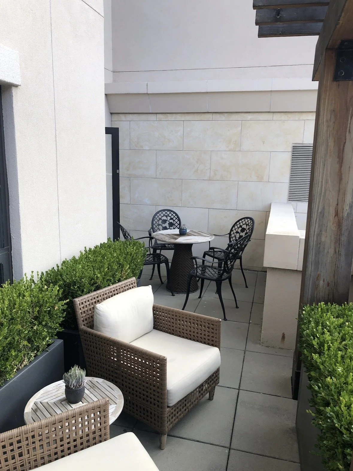 A small outdoor patio with a wicker armchair and a small round table with a potted plant, surrounded by green bushes. In the background, there is a black metal table with four matching chairs and a box or ledge against the wall. The patio has a tiled floor and beige stone walls with a vent.