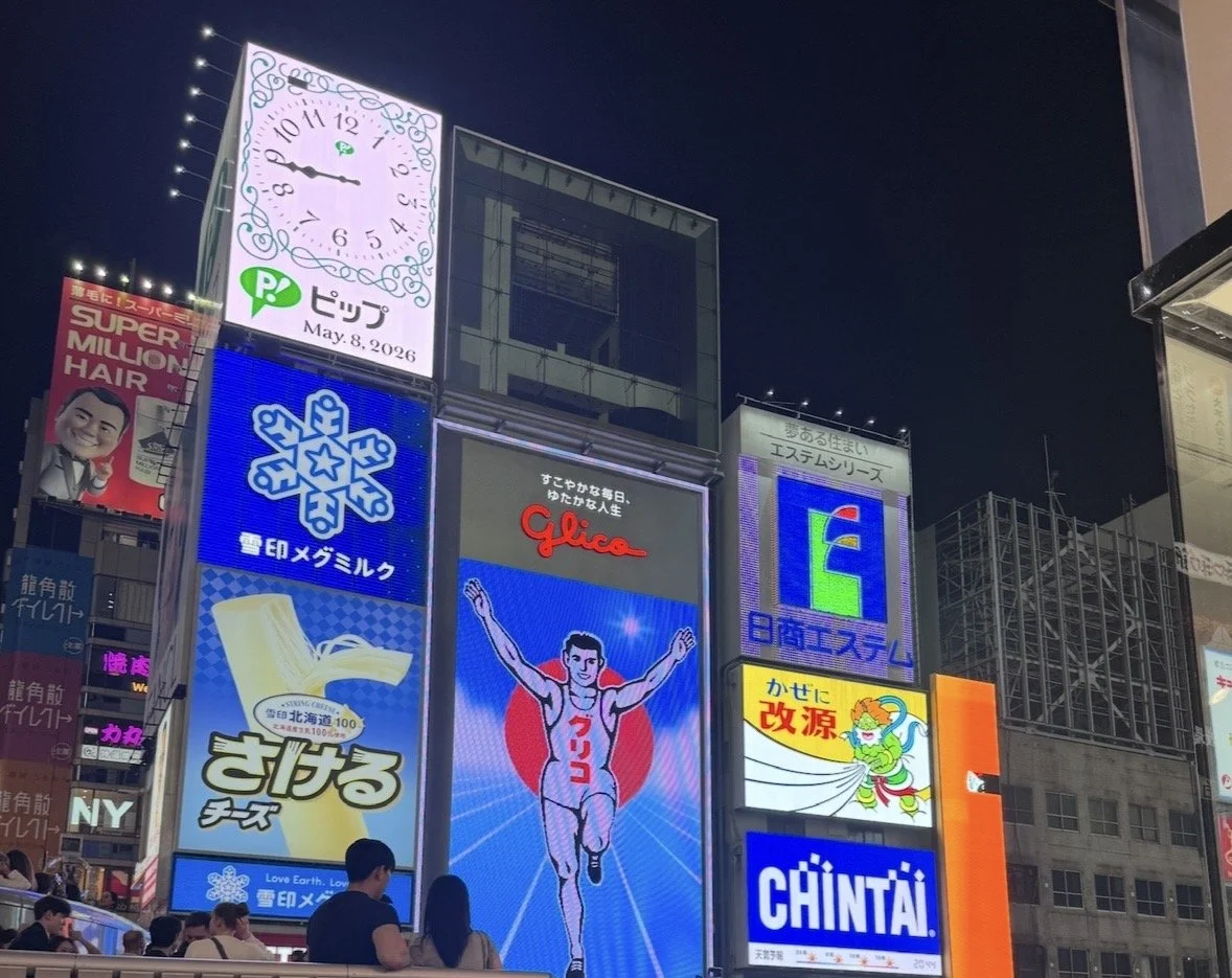 Nighttime cityscape with illuminated billboards and advertisements, including a clock, a runner, and various colorful signs in a busy urban area.