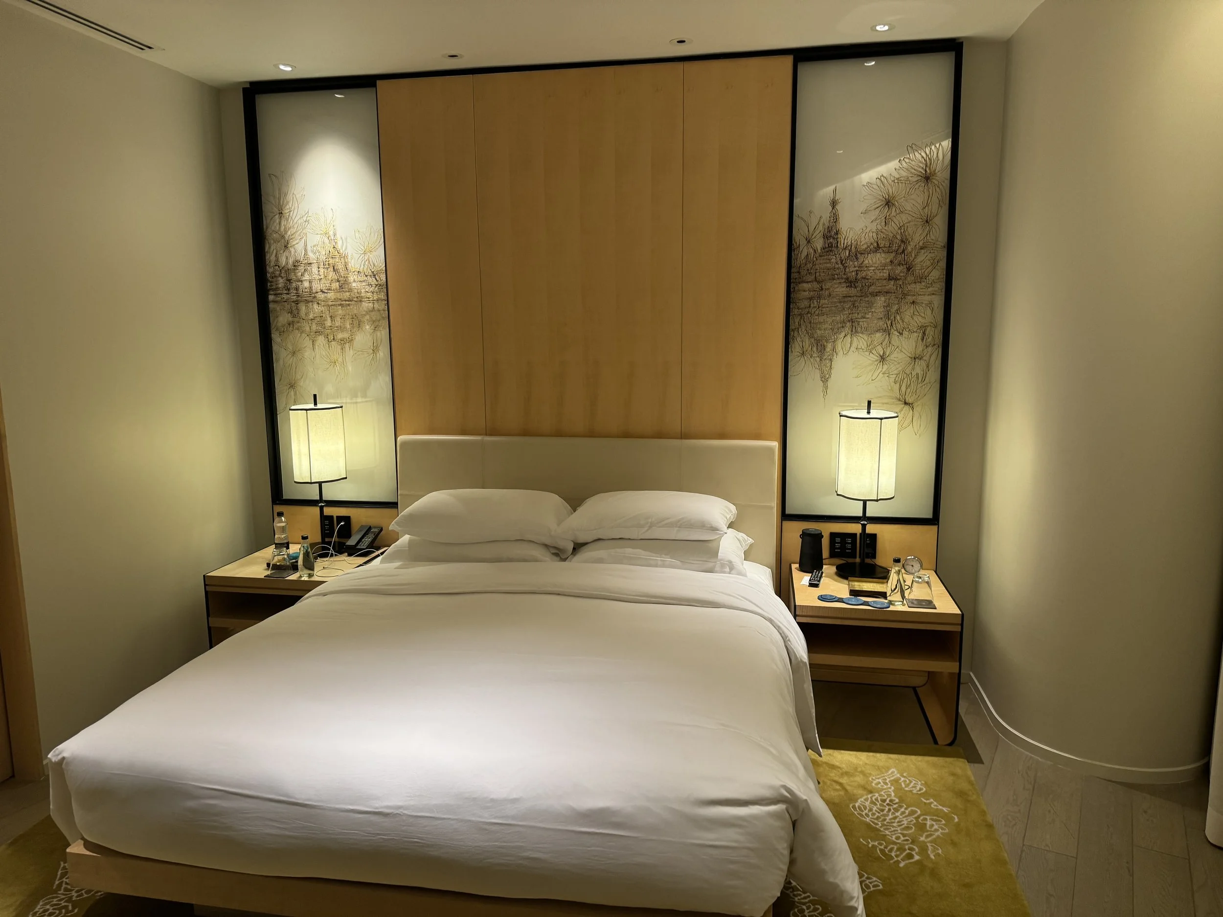A hotel room with two beds, artificial lighting, bedside lamps, a nightstand with a water bottle, remote controls, and decorative panels with artwork behind the beds.