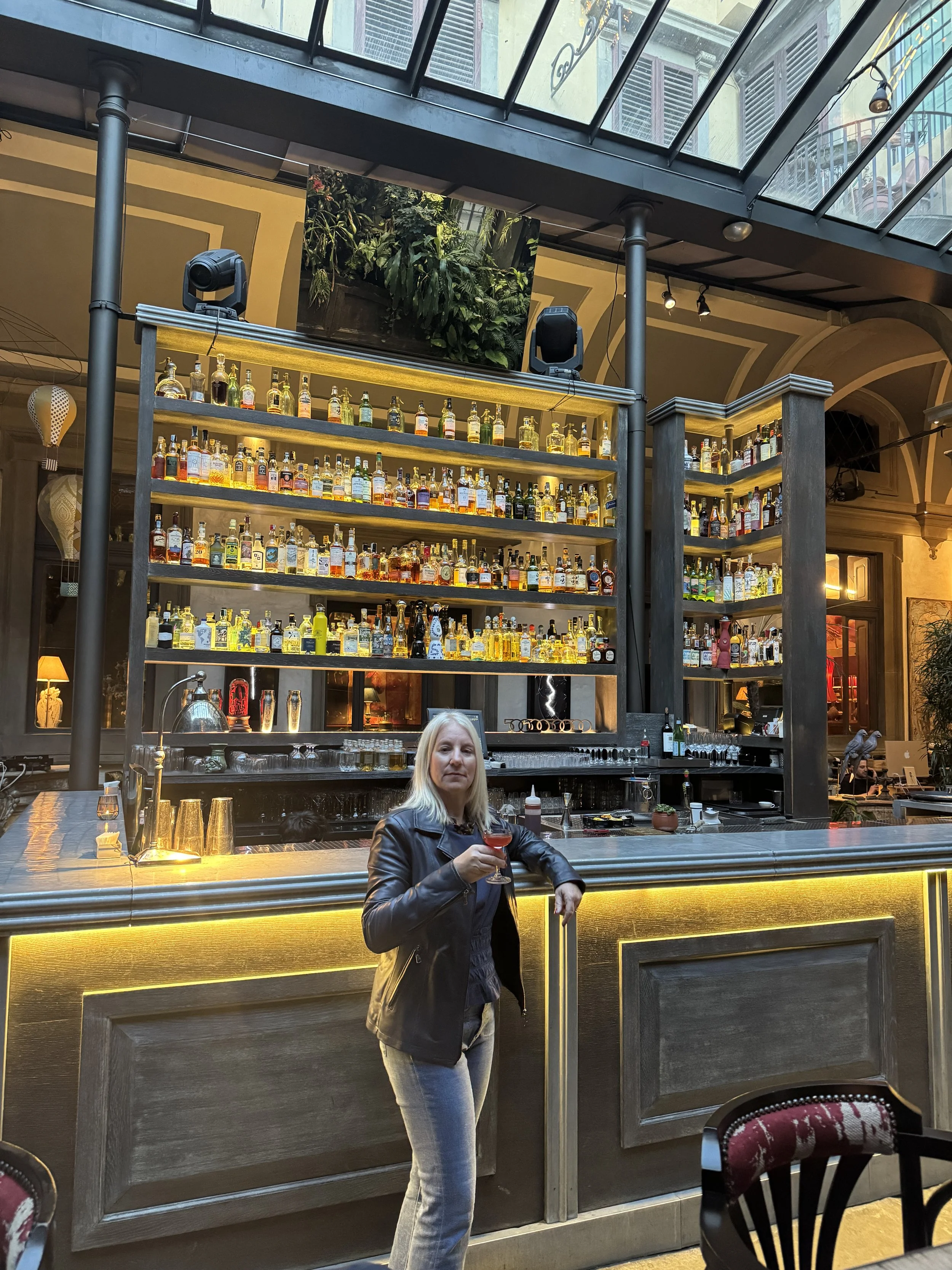 A woman in a leather jacket and jeans standing in front of a bar with a glass of red drink, inside a restaurant or bar with a large liquor shelf, high ceilings, and modern decor.