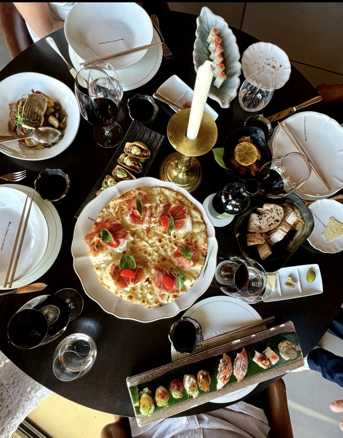A top-down view of a table set for a meal with various dishes including pizza, sushi, bread, and appetizers, along with glasses of red wine, water, and a candle centerpiece.