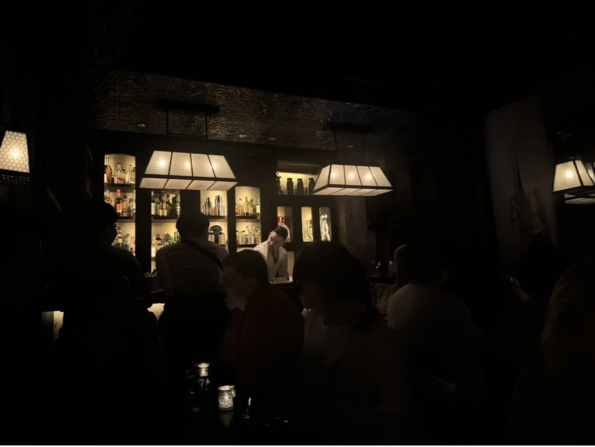 A dimly lit bar with shelves of liquor bottles, a bartender, and patrons sitting at the counter.