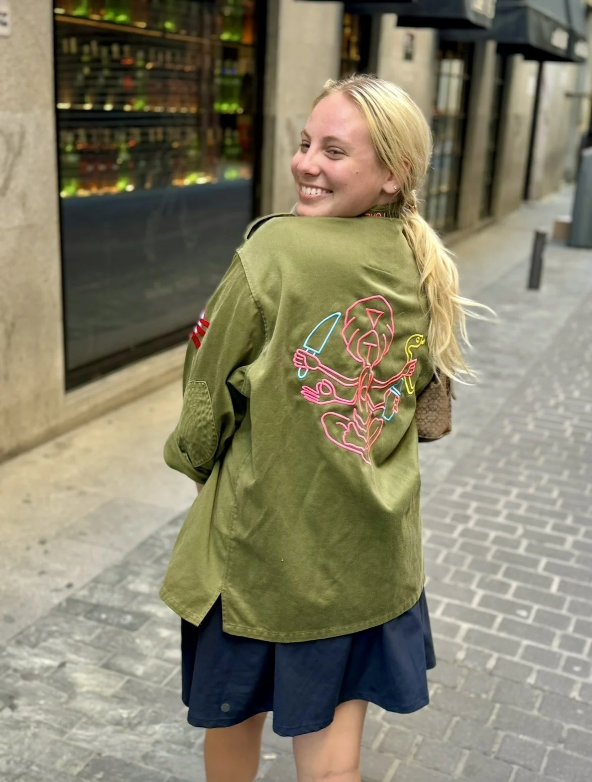 A young woman with blonde hair smiling and turning to her right, wearing an olive green jacket with a neon pink and blue embroidered design of a cartoon figure holding a knife on the back, and a dark skirt, walking on a city sidewalk at night.