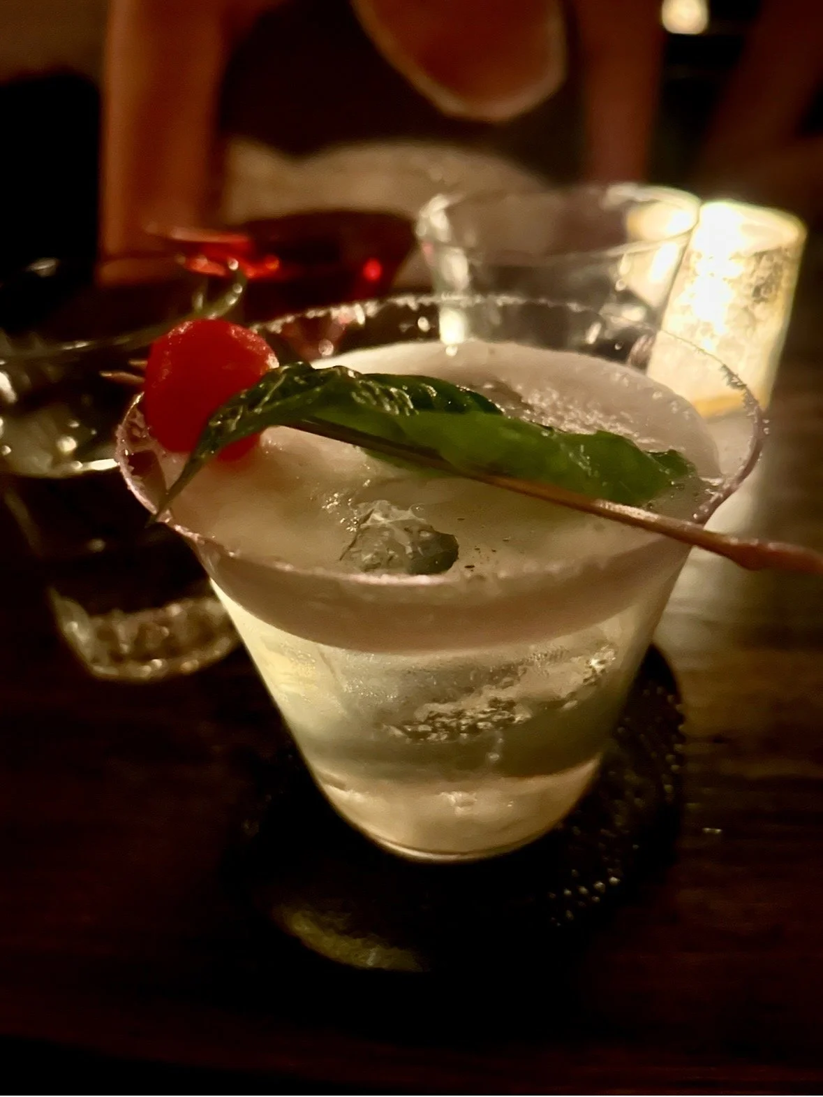 A cocktail in a martini glass garnished with a cherry and a green leaf, served on a dark wooden table at a dimly lit bar or restaurant.