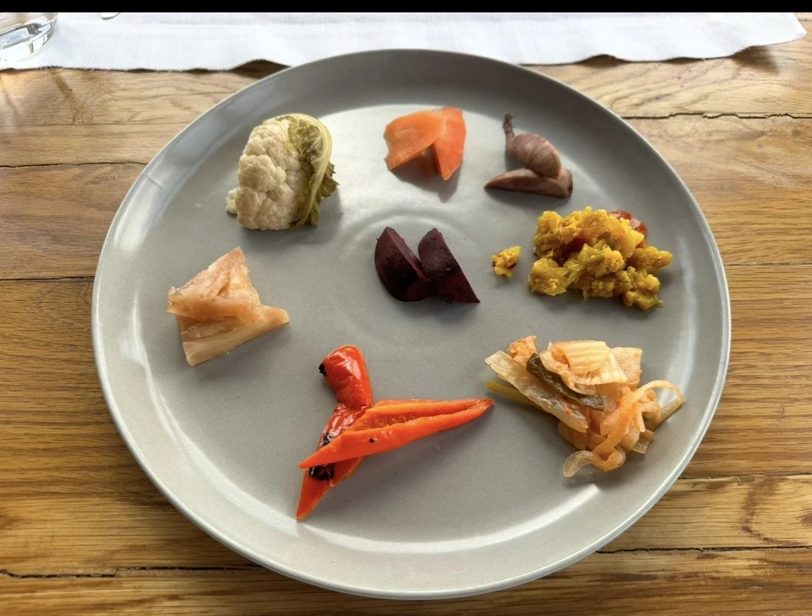 A light gray plate with various vegetables, including cauliflower, carrot, beet, sweet potato, mustard greens, pickled vegetables, and roasted red pepper, placed on a wooden table.