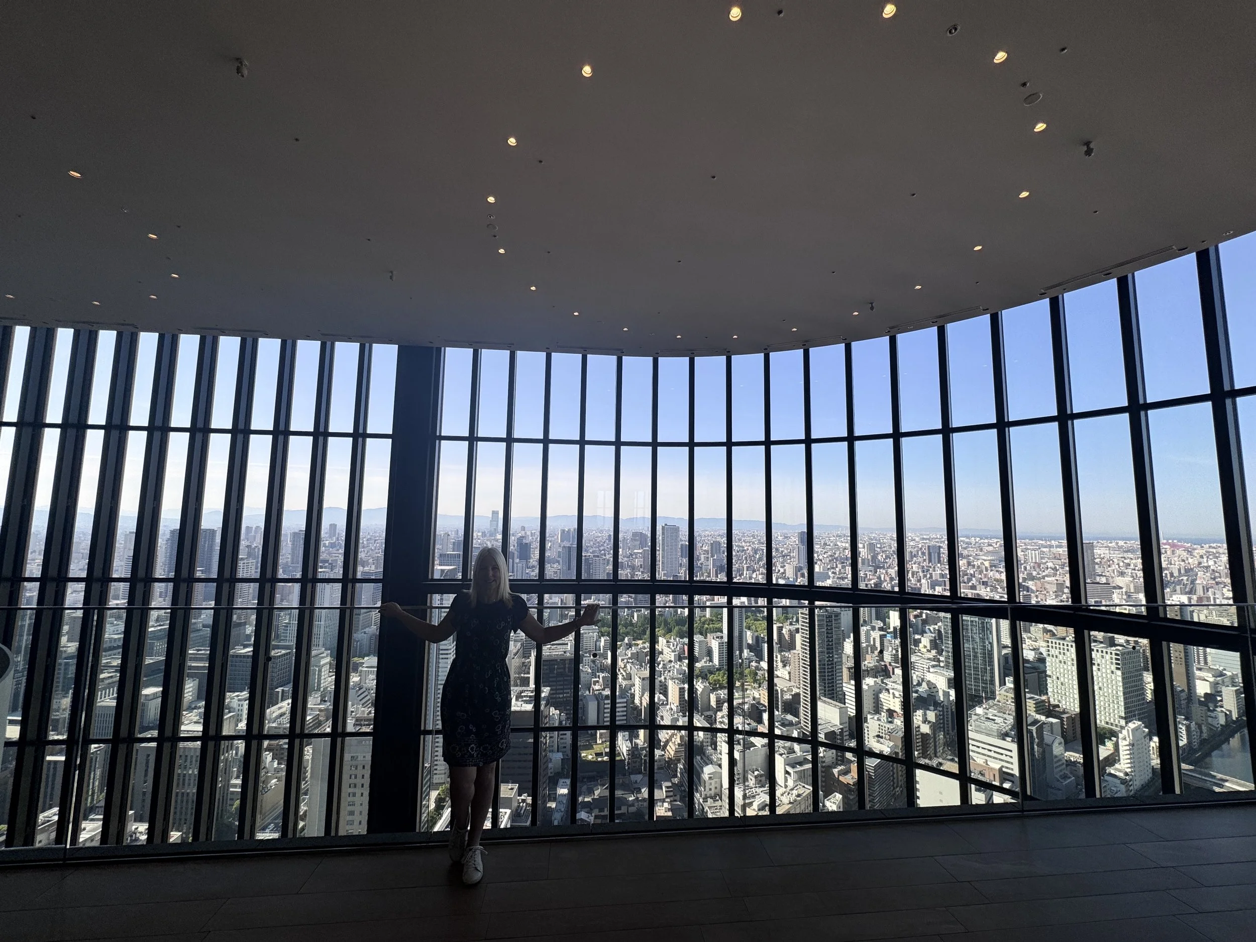 A woman standing inside a modern skyscraper with large glass windows, overlooking a cityscape with numerous high-rise buildings below and mountains in the distance.