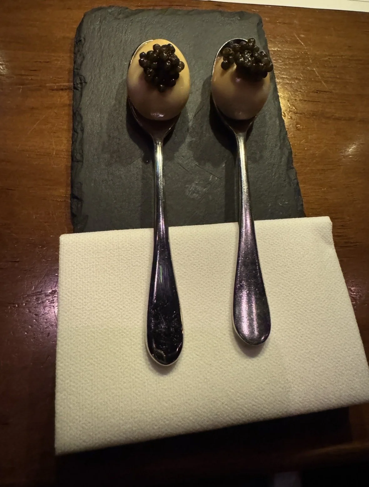 Two spoons with chocolate mousse topped with black tapioca pearls, placed on a black slate serving plate on a wooden table.