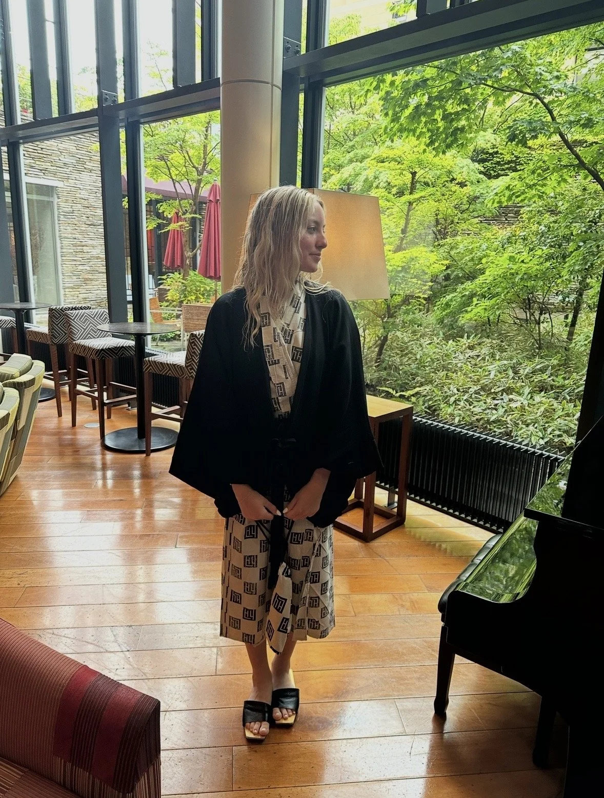 A woman with wavy blonde hair standing indoors near large windows with greenery outside, wearing a patterned dress, black jacket, and black sandals.