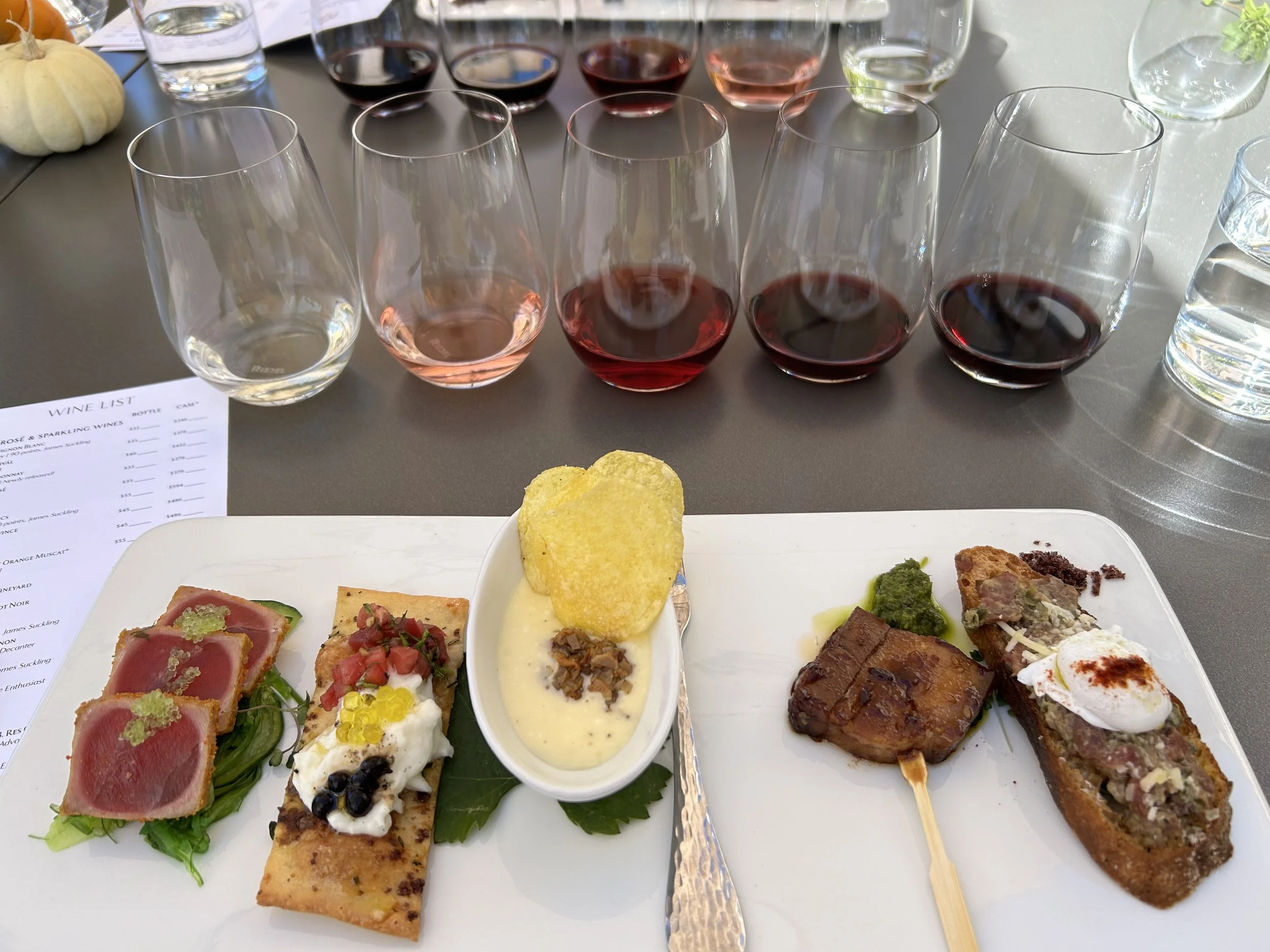A white rectangular plate with assorted appetizers, including seared tuna with green garnish, a slice of quiche topped with faro and blueberries, a small bowl of creamy sauce with a browned topping, grilled meat skewers, and a small section of a fried item. In the background, a sheet of paper with a wine list, several glasses of wine and water, and a white pumpkin are visible.