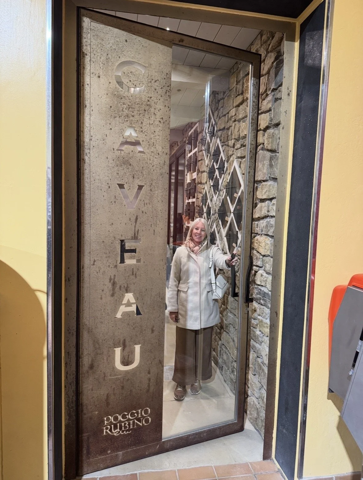 A smiling woman standing inside a glass door with a steel frame. The door has the word 'CASA' vertically cut into it, and the name 'POGGIO RUBINO' at the bottom. Behind her, the interior wall is decorated with a geometric design of white lines and faux stone.