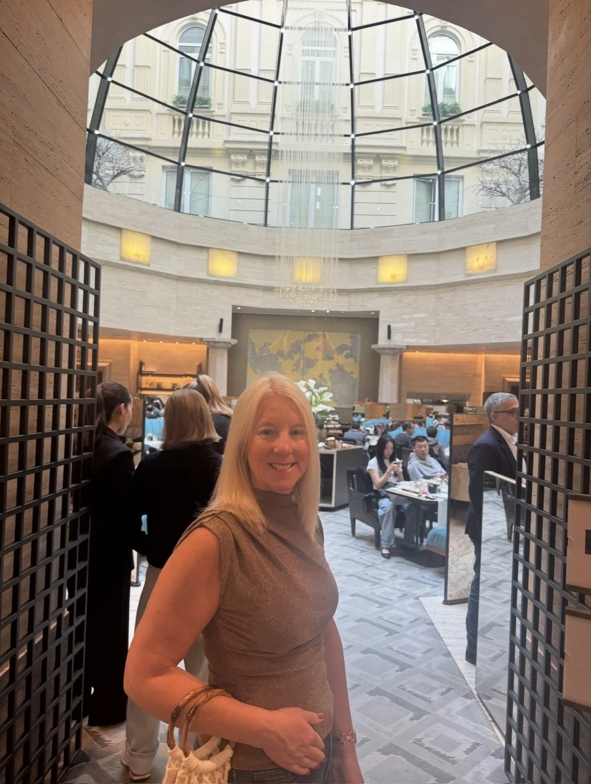 A woman with blonde hair smiling at the camera, standing in a restaurant or hotel lobby with a glass ceiling and several people dining in the background.