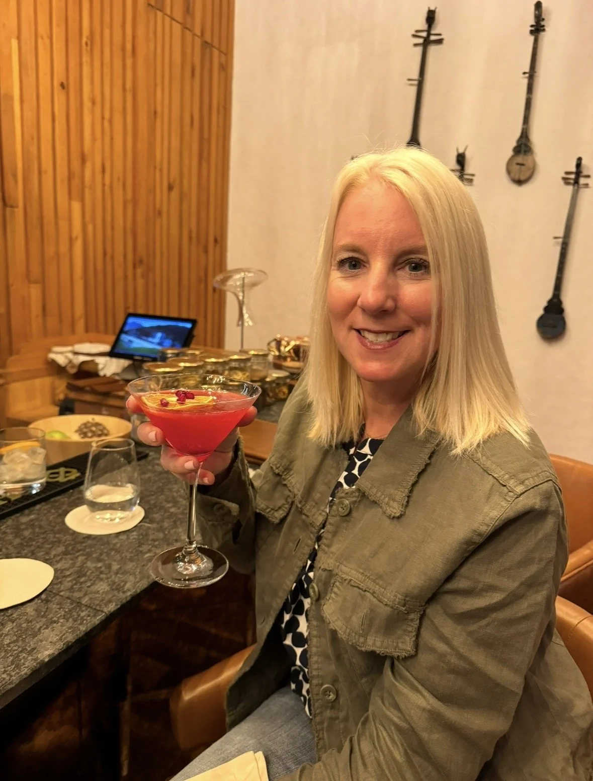 A smiling woman with blonde hair holding a red cocktail in a martini glass in a restaurant setting.