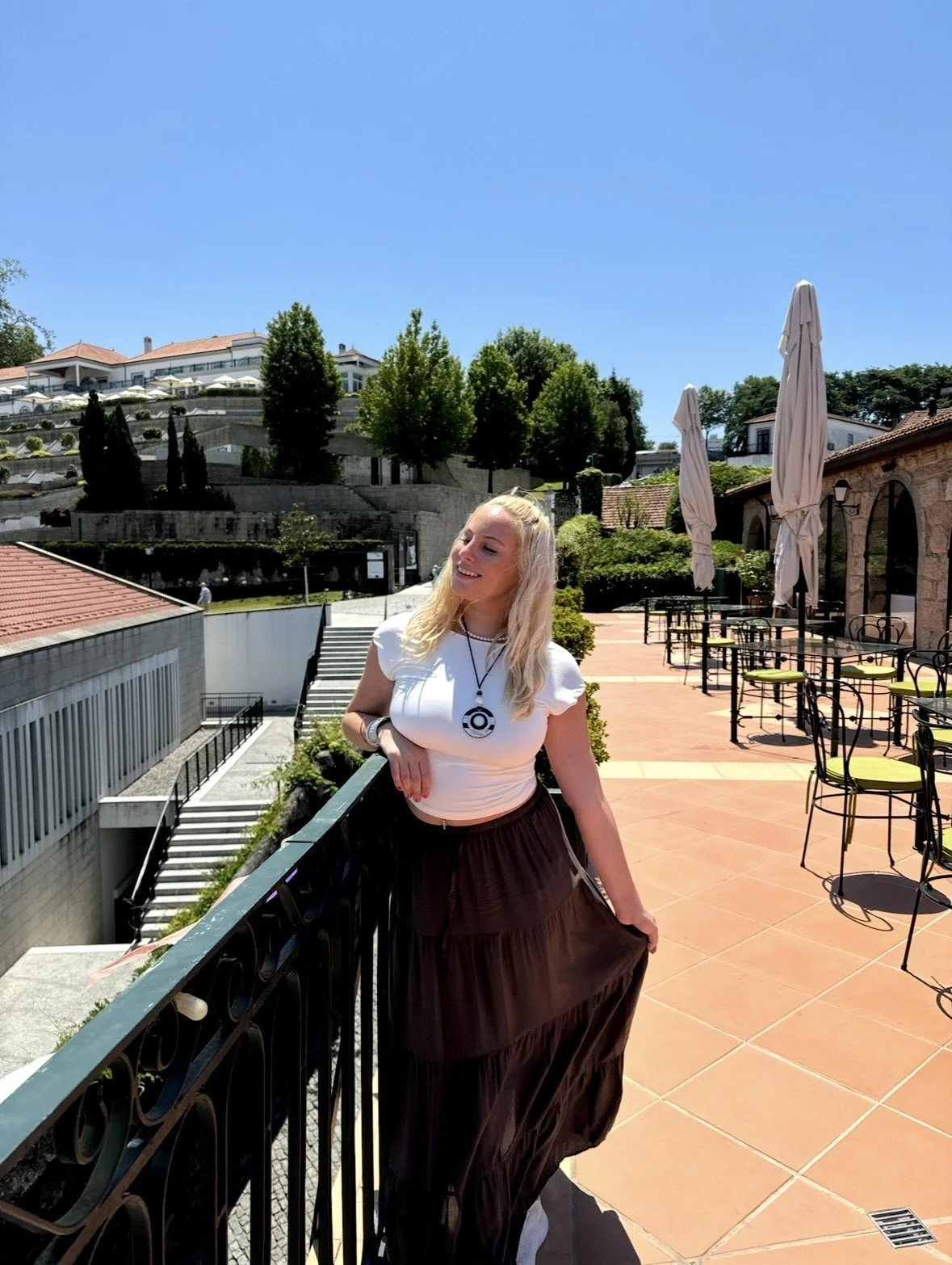 A woman with blonde hair, wearing a white top and long dark skirt, leaning on a black railing on a sunny outdoor terrace with green trees, umbrellas, and buildings in the background.