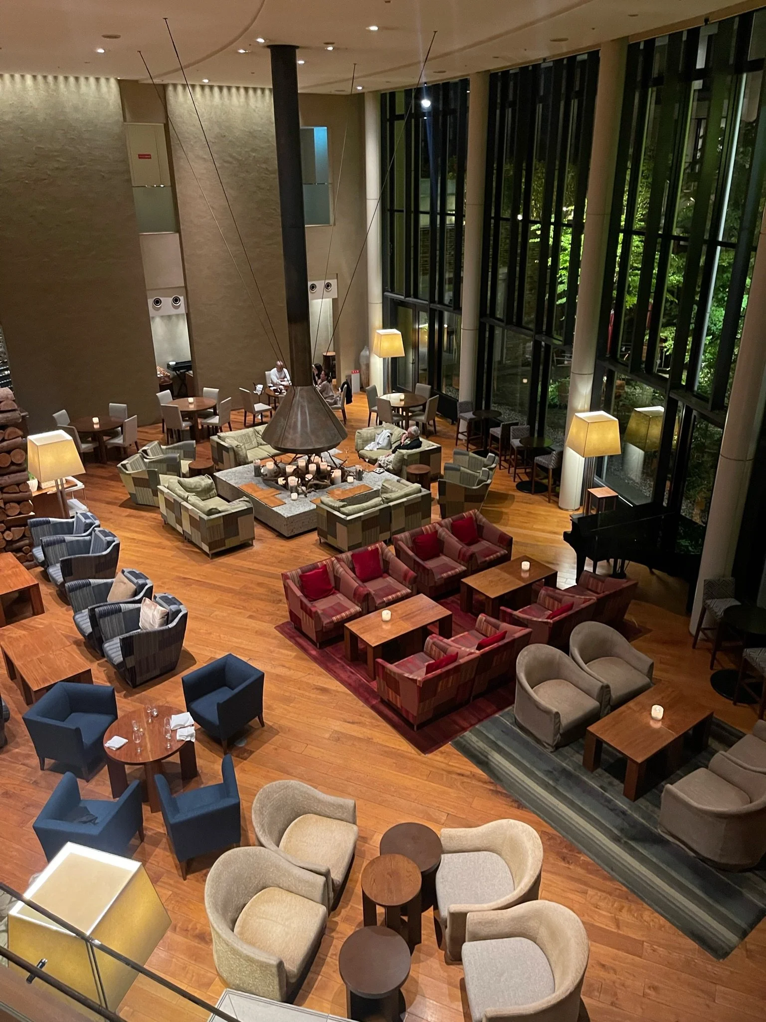 A spacious hotel lobby with high ceilings, large windows, and various seating arrangements including armchairs, sofas, and tables. There is a grand piano in the foreground, and the area is illuminated with warm lighting from lamps and the ceiling. Outside visible through the windows are trees and outdoor lighting.