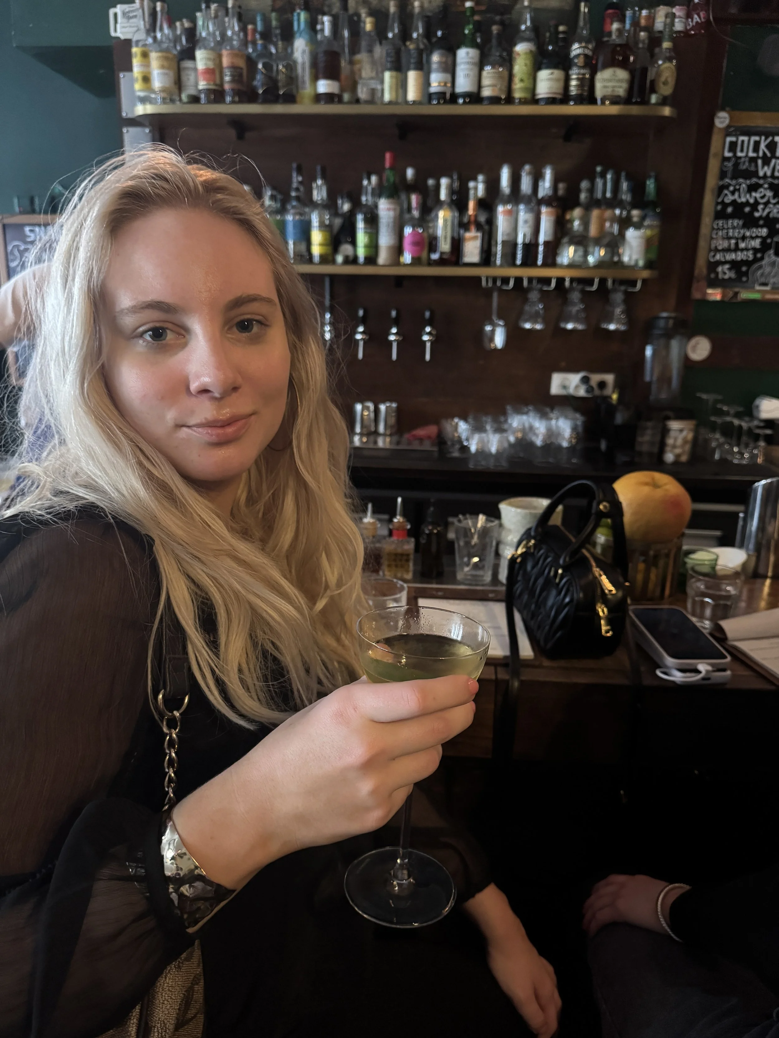 A young woman with blonde hair holding a glass of white wine at a bar, with shelves of liquor bottles in the background.