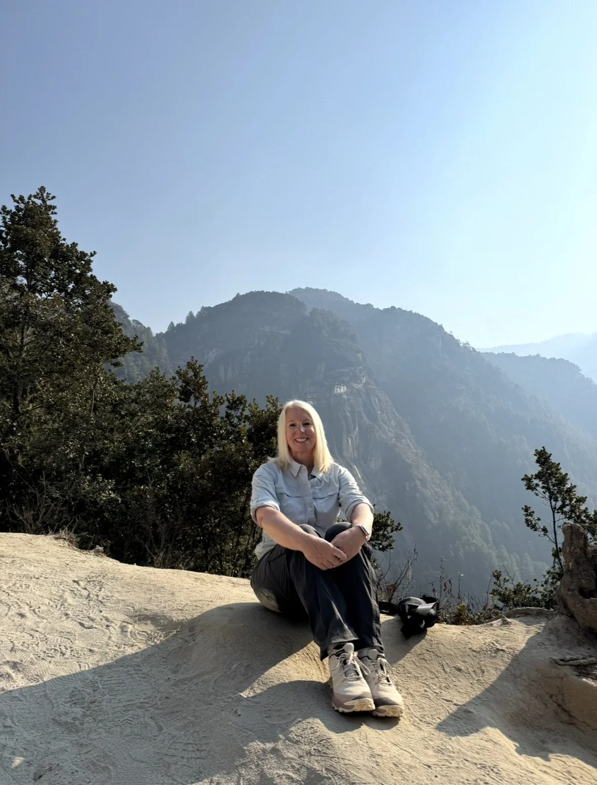 A woman sitting on a rock in a mountainous area with trees and a clear blue sky.