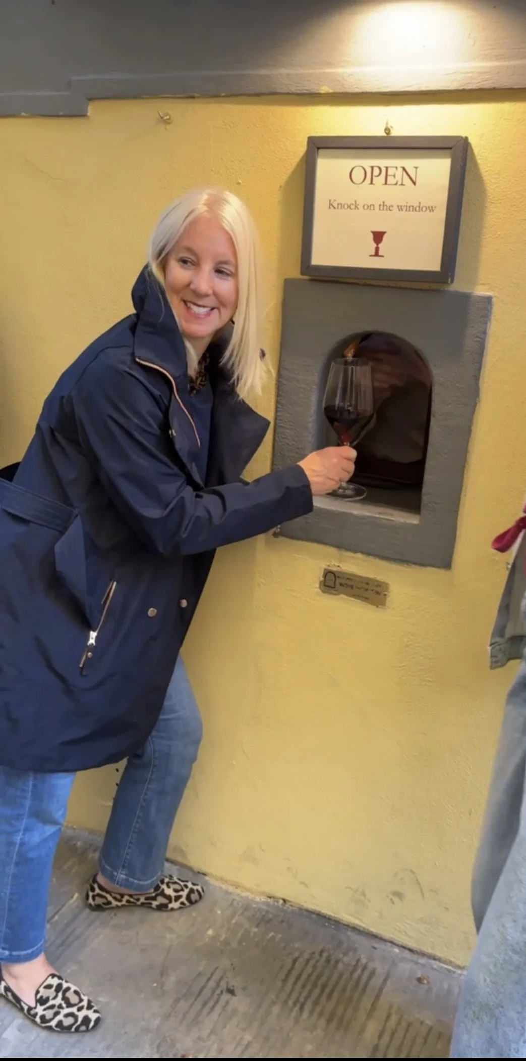 Woman with blonde hair, wearing a navy jacket and leopard-print shoes, holding a glass of red wine at an outdoor window with a sign that says 'Open, Knock on the window'.