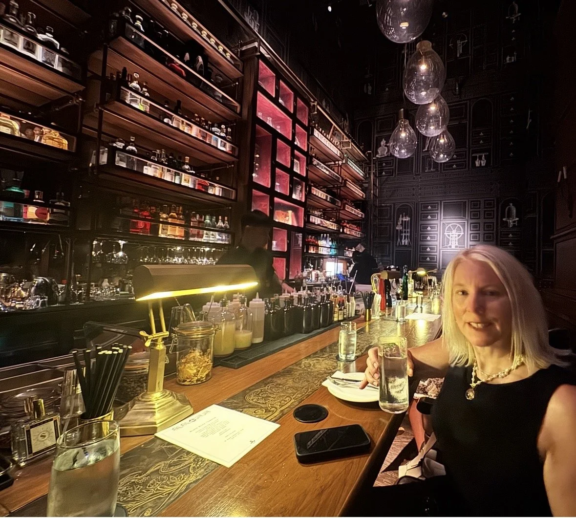 A woman sitting at a bar holding a water glass, smiling, with a dark, elegantly decorated bar interior in the background featuring shelves of bottles and glasses, and pendant lights overhead.