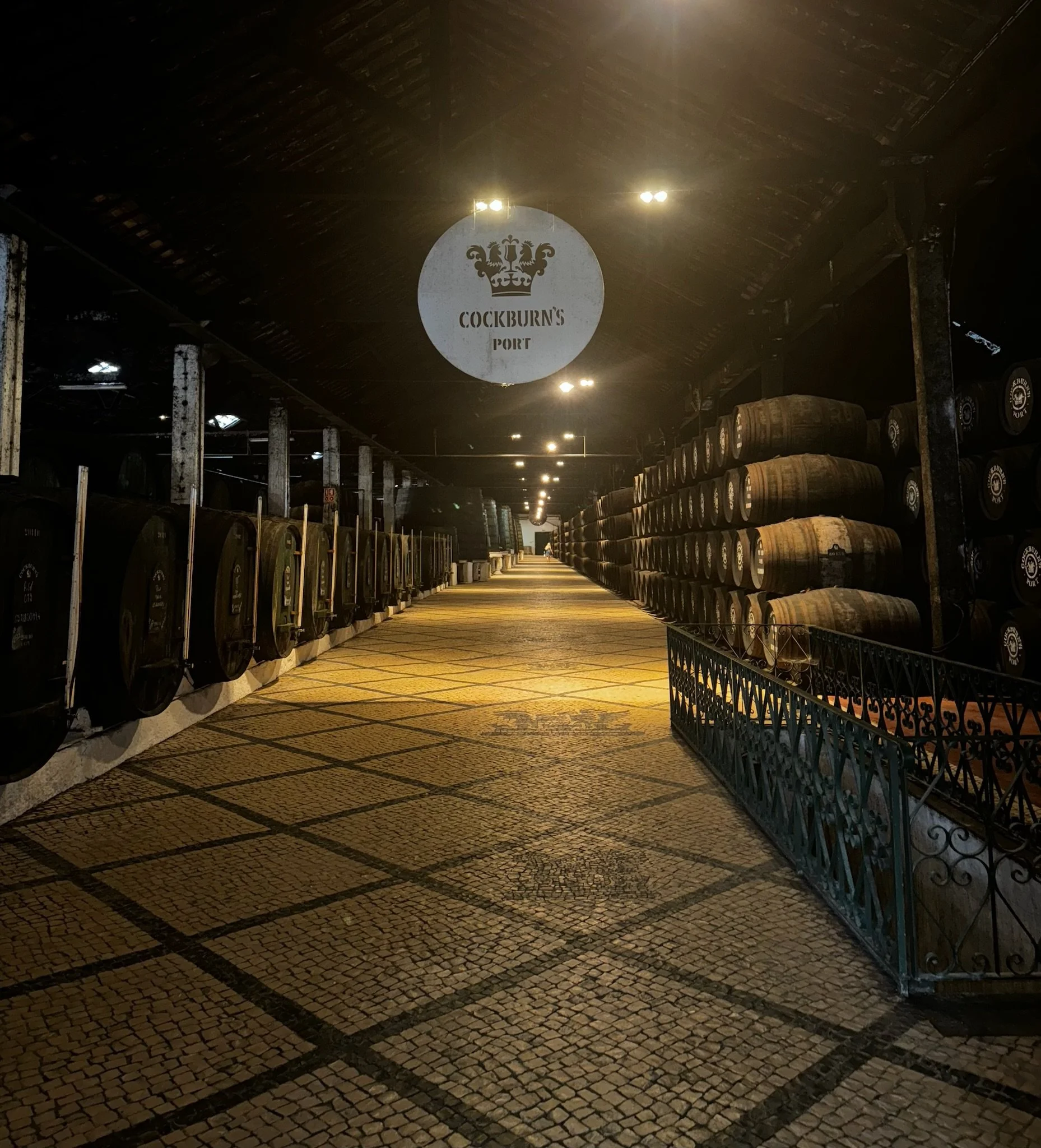 Interior of a whisky cellar with barrels stacked on racks and a sign reading 'Cockburn's Port' hanging from the ceiling.