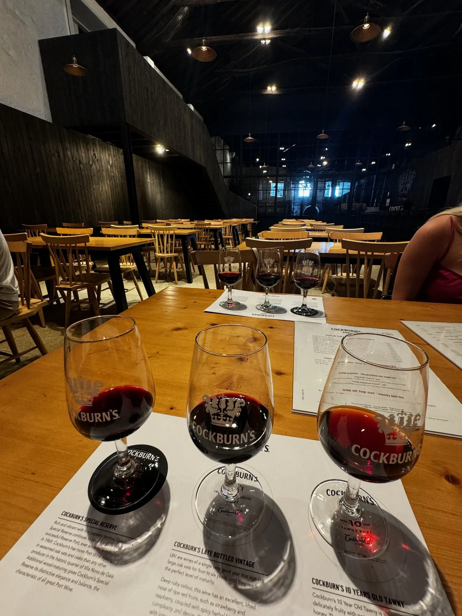 Three glasses of red wine on a wooden table with printed menus underneath, inside a modern restaurant with wooden chairs and dim lighting.