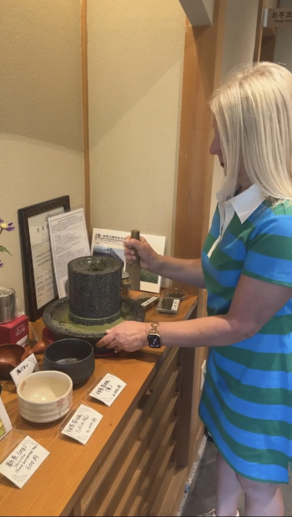 A woman with blonde hair wearing a blue and green striped shirt and a watch on her left wrist is serving food at a counter. There are various bowls and signs with Japanese writing on the counter, and she is in the process of pouring or serving something into a bowl.