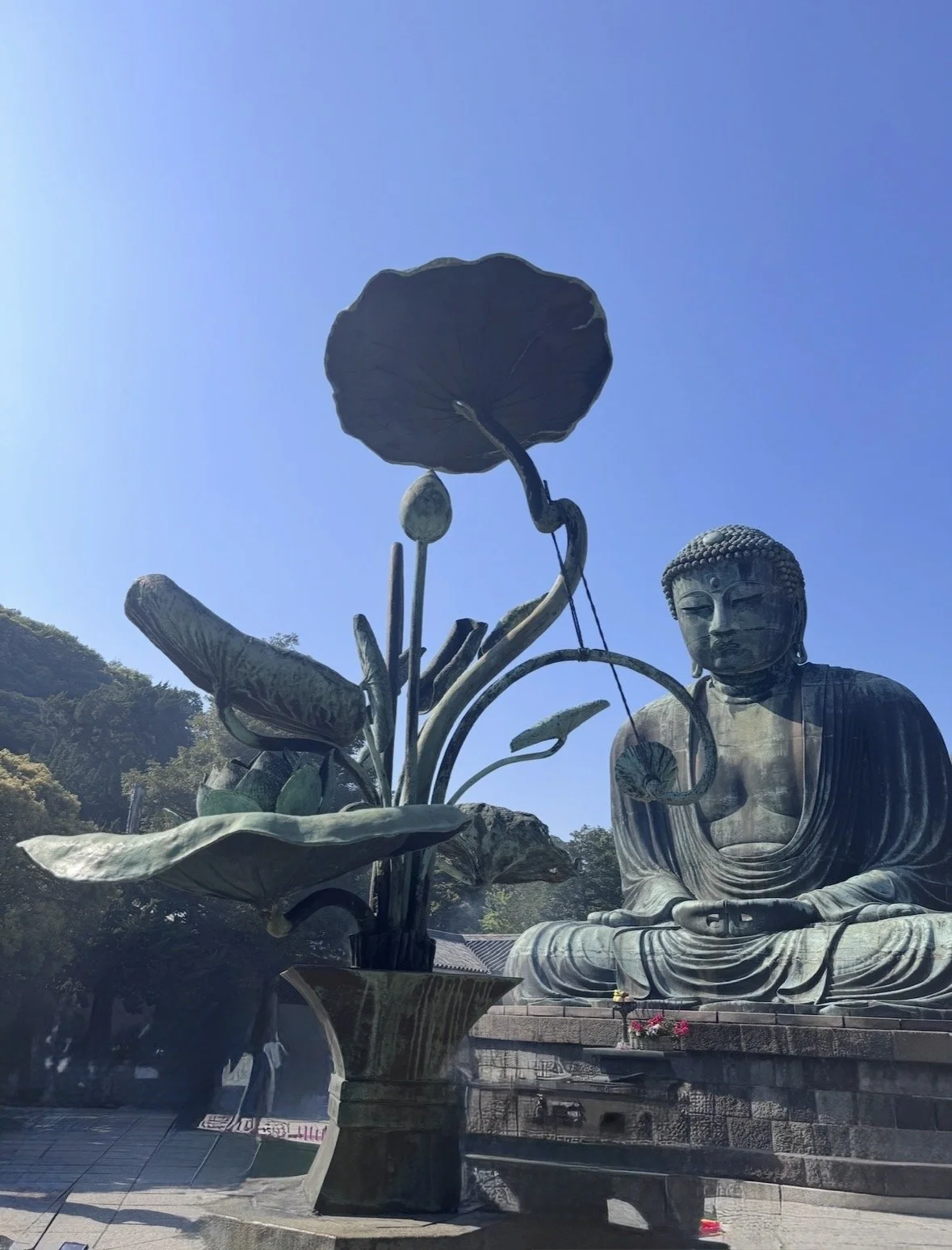 Large outdoor statue of Buddha sitting in a meditative pose next to a big sculpture of a lotus flower in a garden setting.