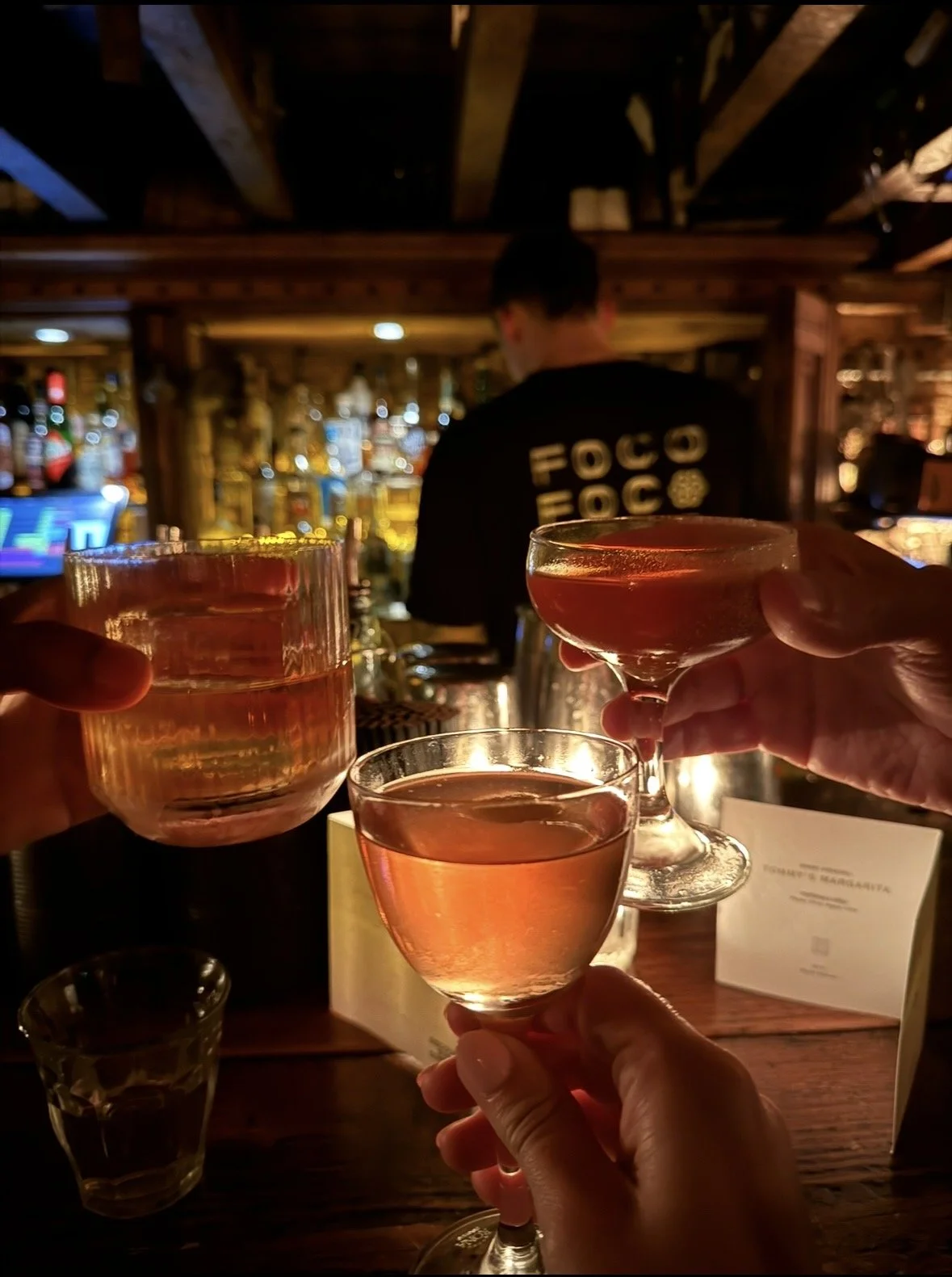 Three people at a bar raising glasses for a toast, with a bartender and bar in the background.