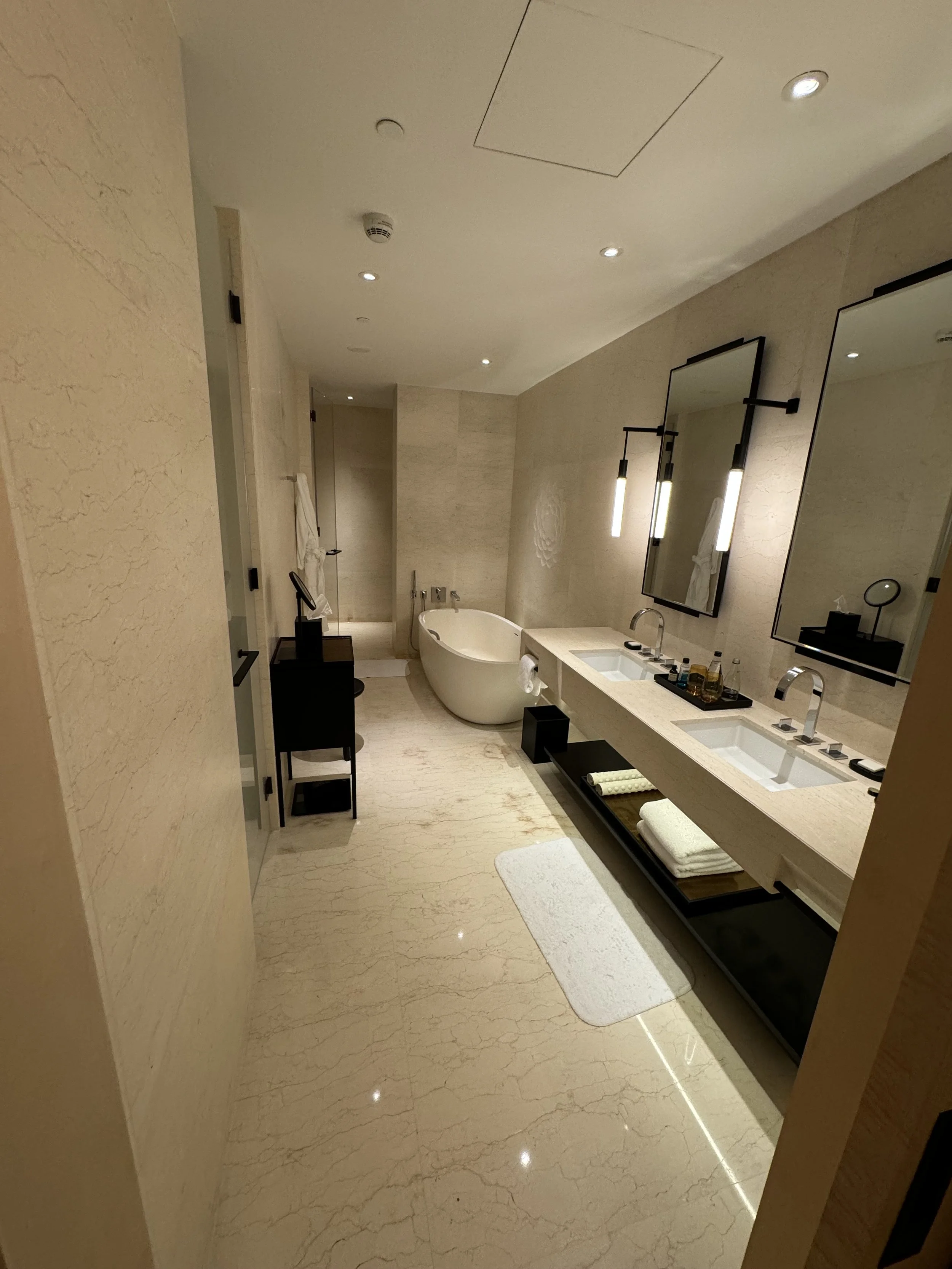 A luxurious hotel bathroom featuring a double sink vanity, large mirrors, a freestanding bathtub, and elegant beige marble floors and walls.