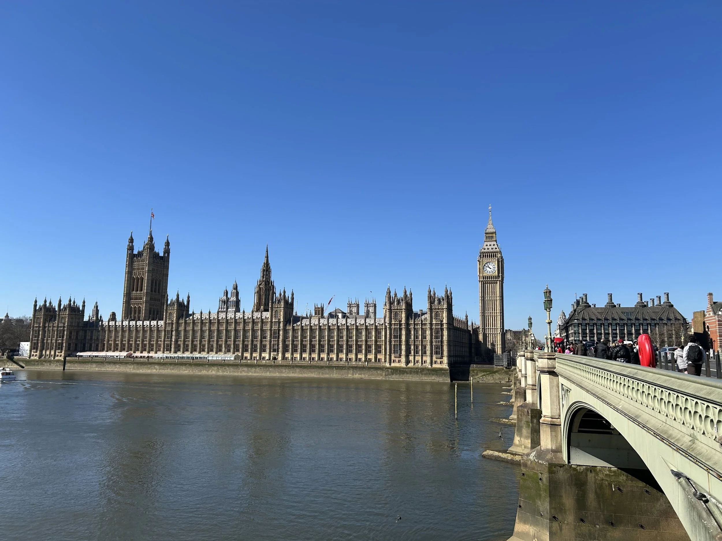 View of the Houses of Parliament and Big Ben in London, England, with a bridge and pedestrians along the River Thames under a clear blue sky.