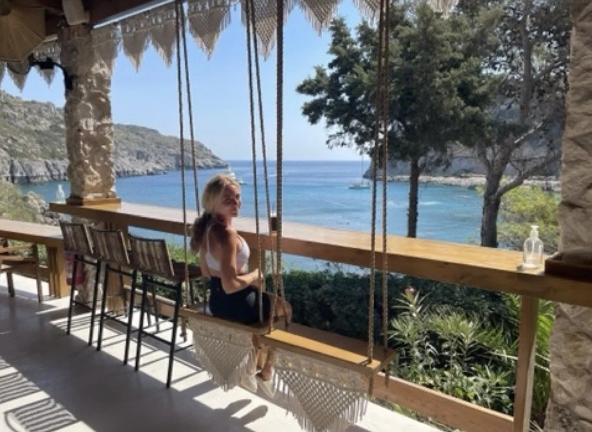A woman sitting on a swing bench on a balcony overlooking the ocean and rocky hills with trees.