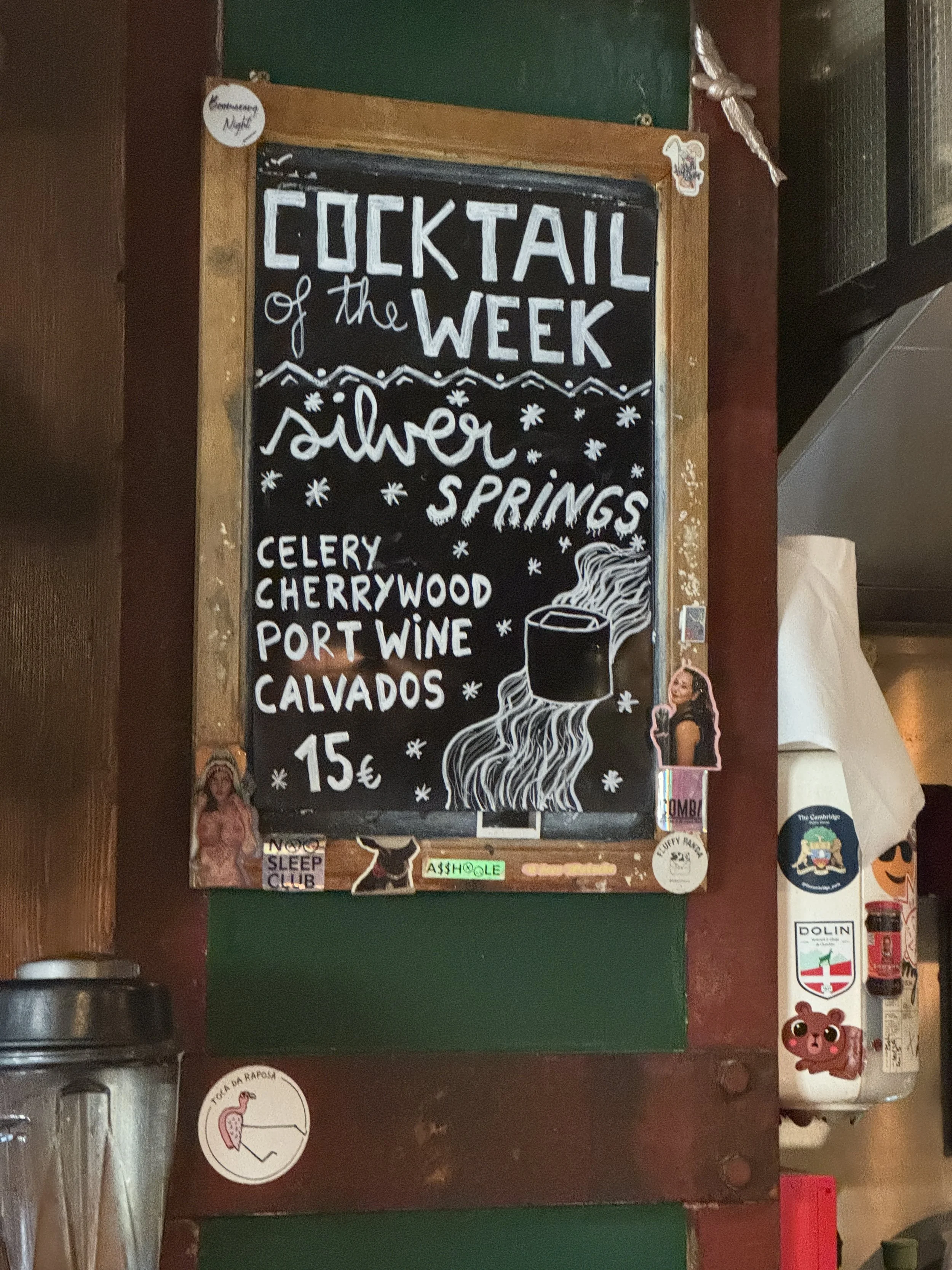 A black chalkboard sign with white handwritten text listing the 'Cocktail of the Week' and its ingredients, decorated with small drawings of stars and a steaming mug, surrounded by various stickers on a wooden surface.