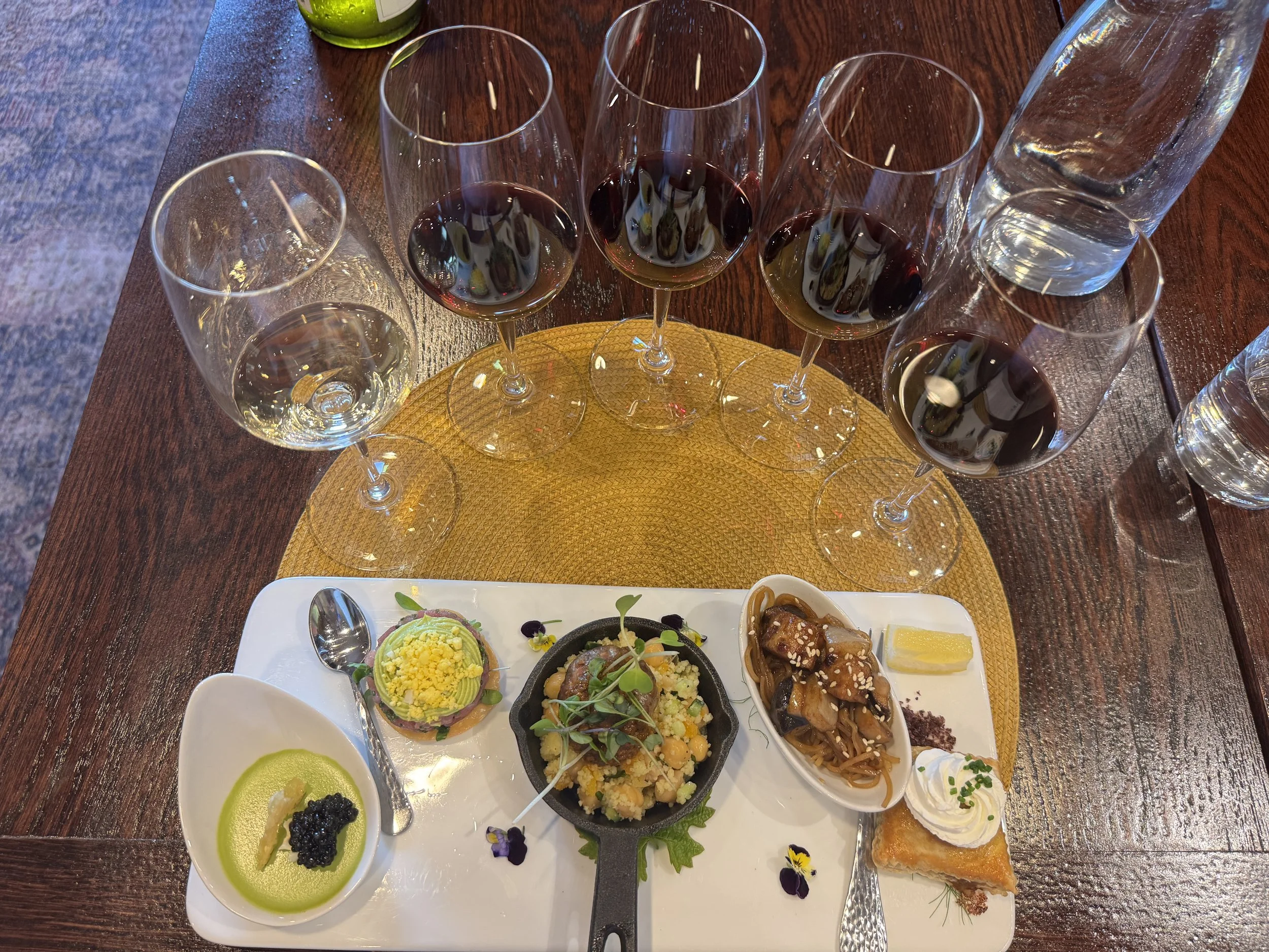 A table with a white platter of assorted Asian-style appetizers, including small bowls of sauces, sushi, and dumplings, along with glasses of red wine and water.