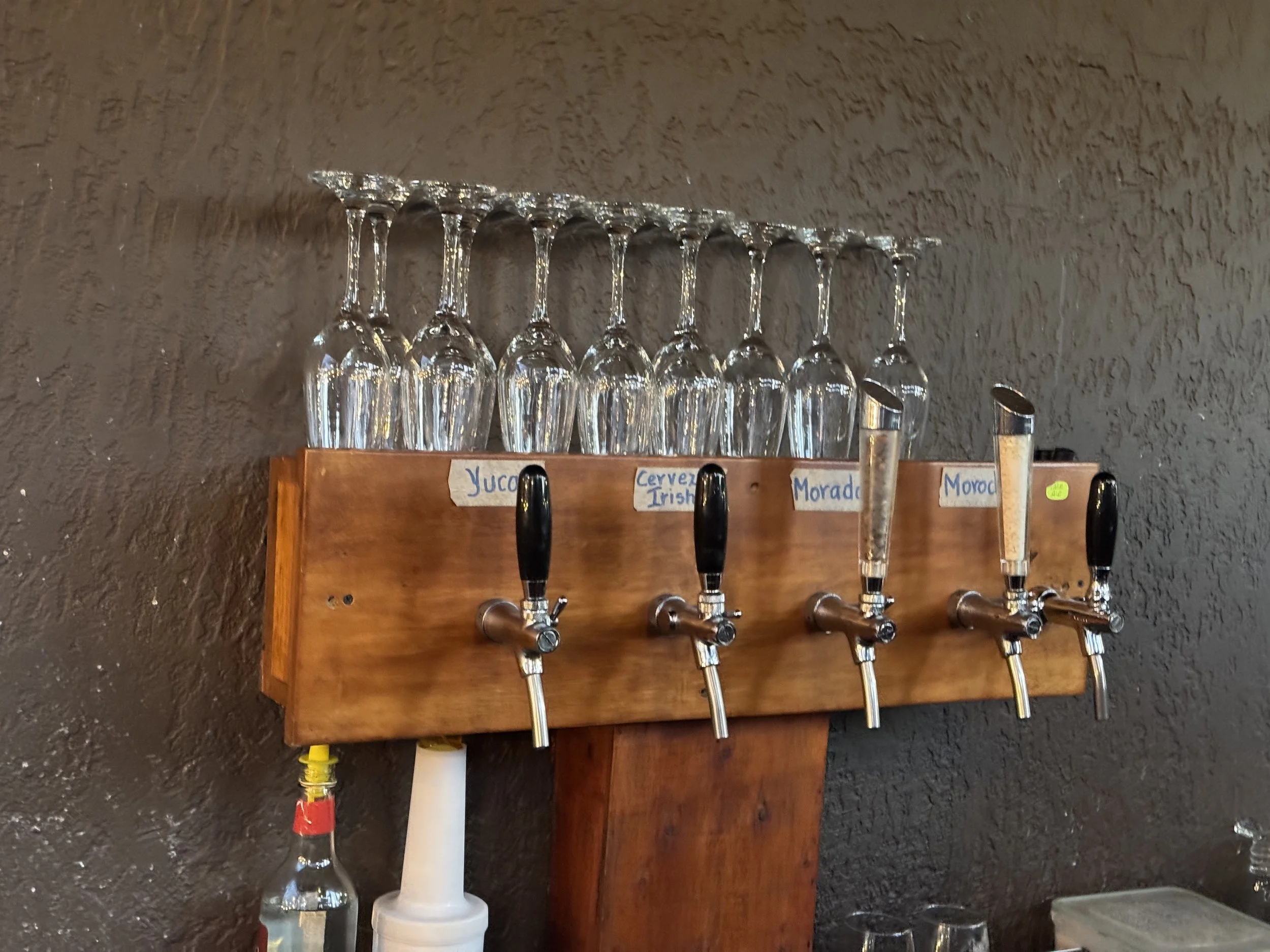 A wooden bar top with several beer taps and a row of upside-down wine glasses hanging above, with small handwritten labels for different beer options.