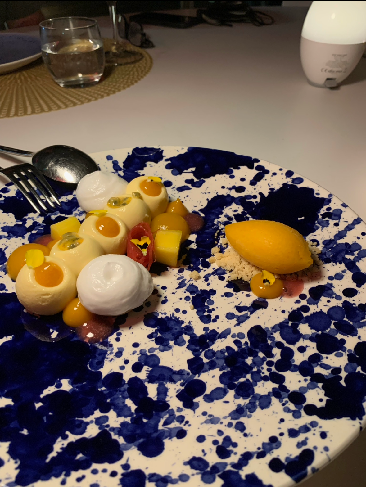 Plate with creative dessert featuring poached eggs, mango cubes, red fruit slices, yellow sauce, spruce tips, and a lemon wedge on blue-speckled plate.
