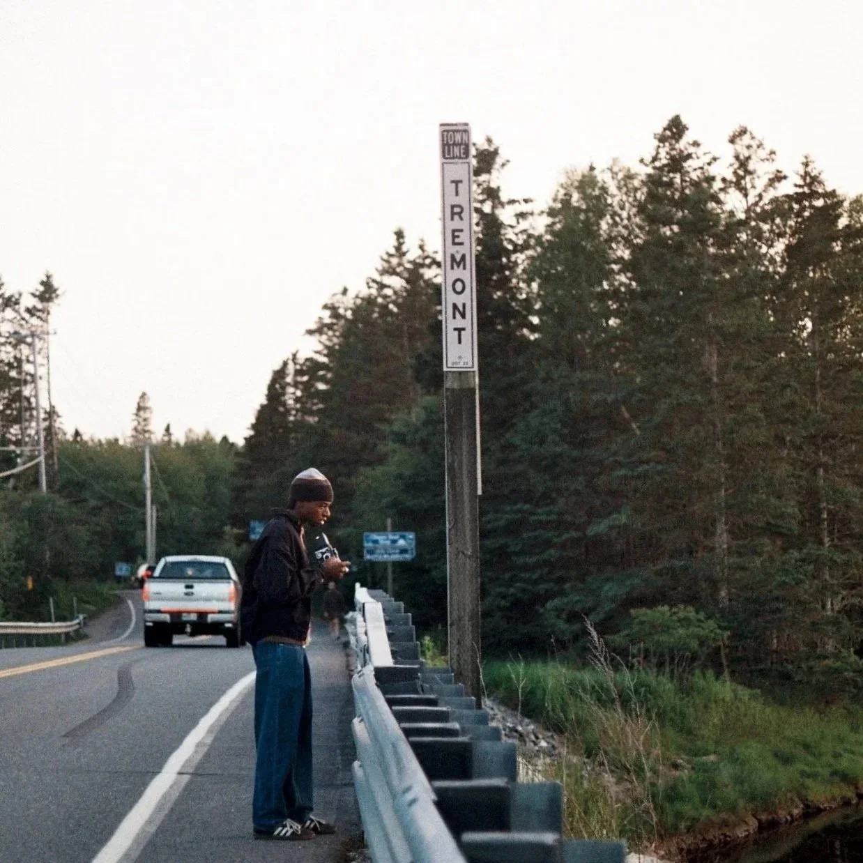 A young person wearing a beanie and jacket standing on the side of a winding road, looking at their phone. There is a white pickup truck ahead on the road, with a guardrail and a signpost that reads "TREMONT" in the background, surrounded by trees.