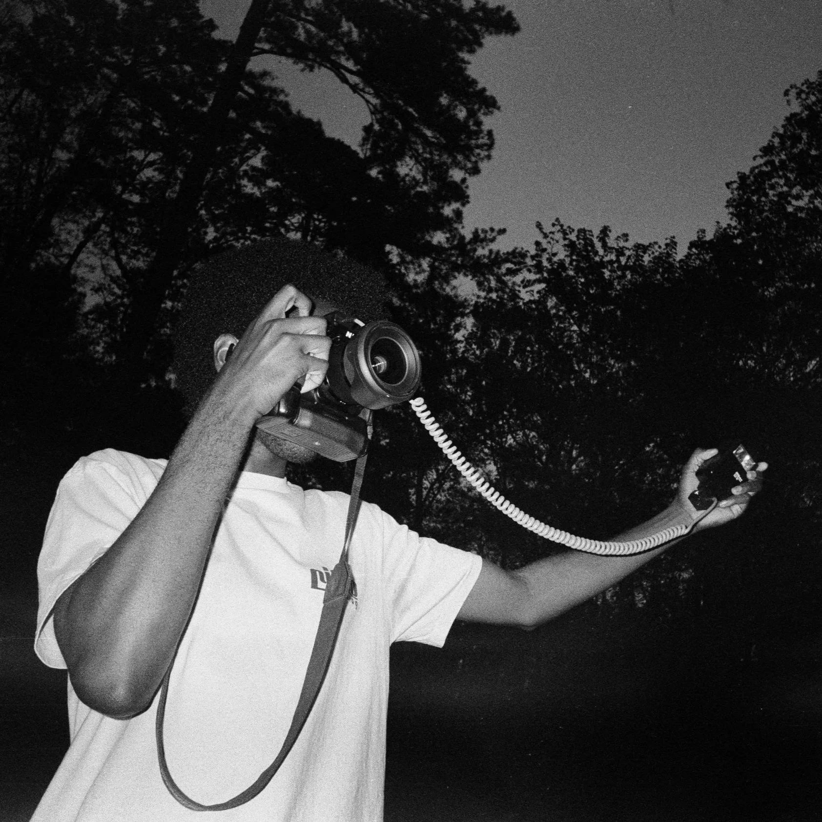 A person wearing a white t-shirt and glasses holding a vintage film camera with a coiled cable outdoors at night, with trees and a star visible in the sky.
