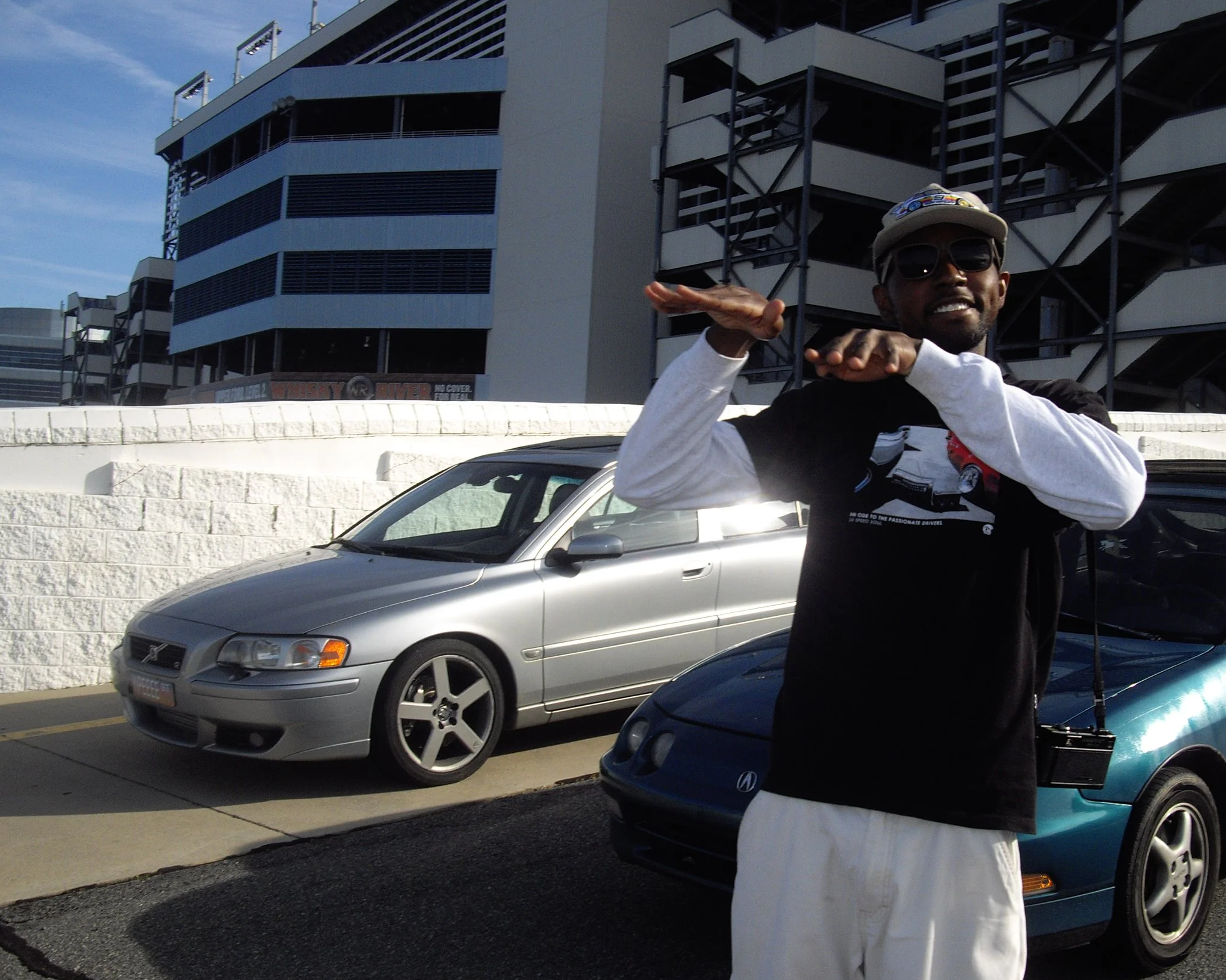Person wearing sunglasses and a cap smiling and gesturing with hand, standing next to parked cars in a parking lot near a modern multi-story building.