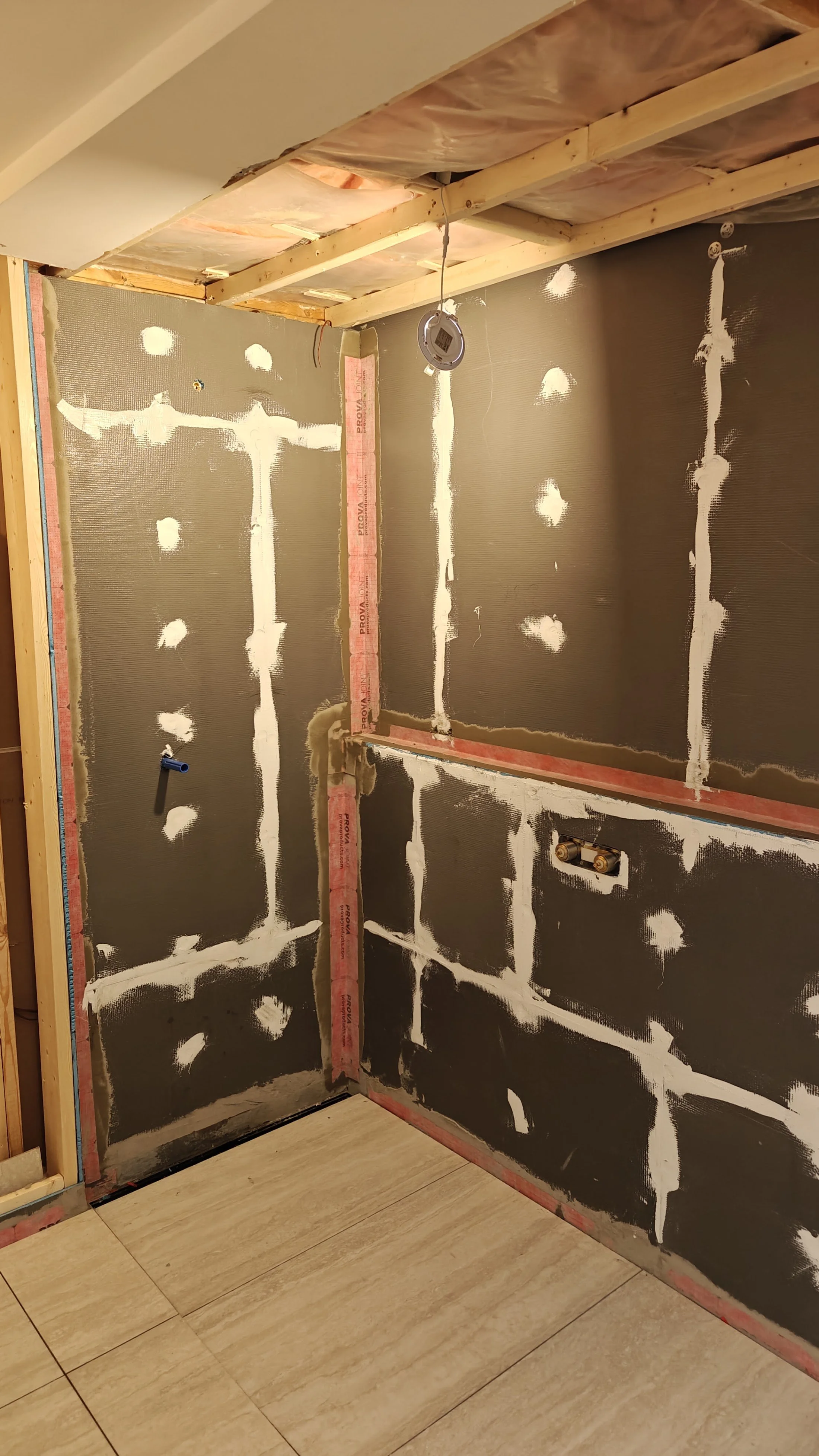 Unfinished bathroom wall with drywall and tape, plumbing visible, and a light fixture hanging from the ceiling, with a tiled floor.