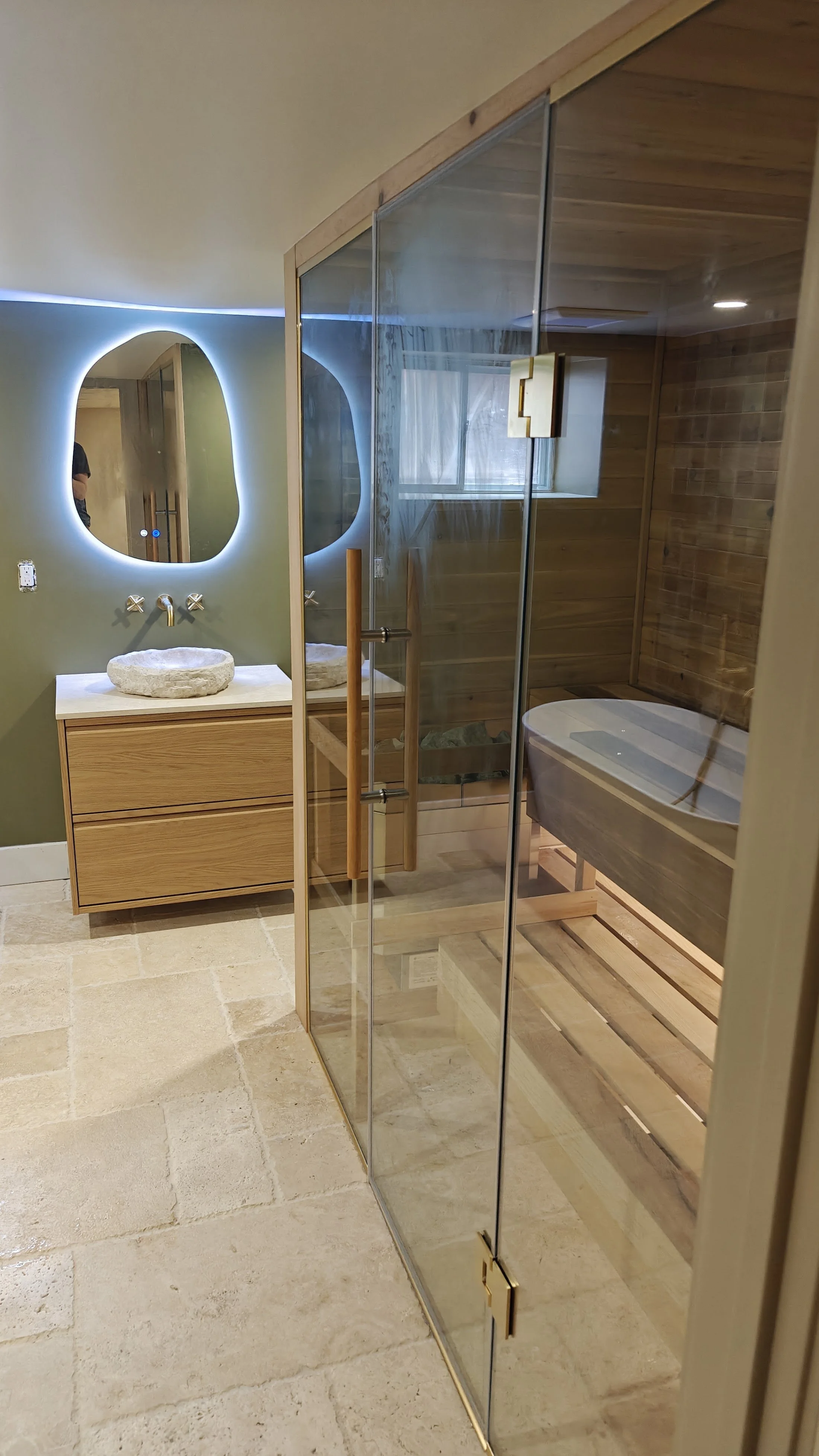 Modern bathroom with green painted wall, oval mirror illuminated from behind, wooden vanity with stone sink, glass shower enclosure with wooden accents, bathtub, and tiled flooring.