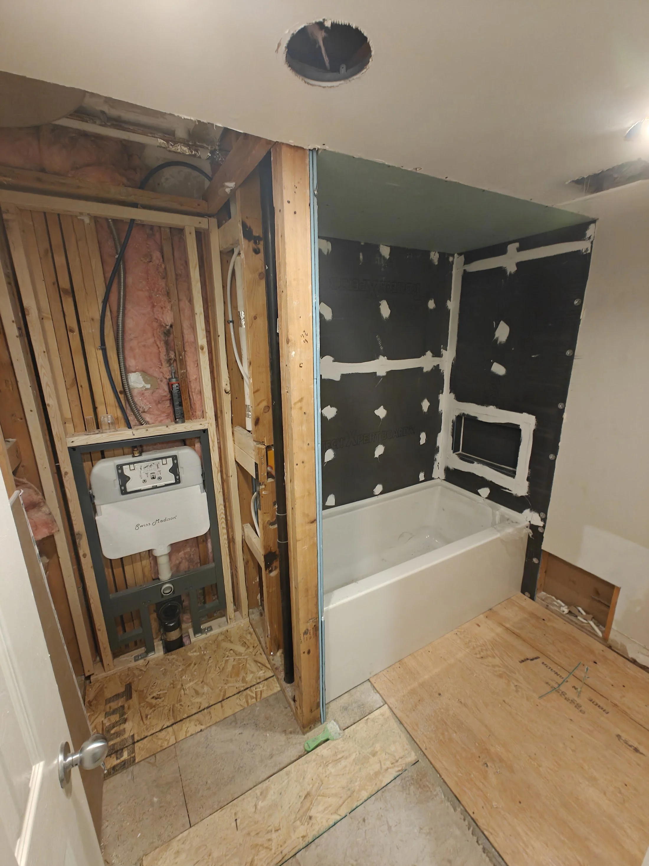 Bathroom renovation under construction with plumbing and framing, drywall partially installed, bathtub in place.