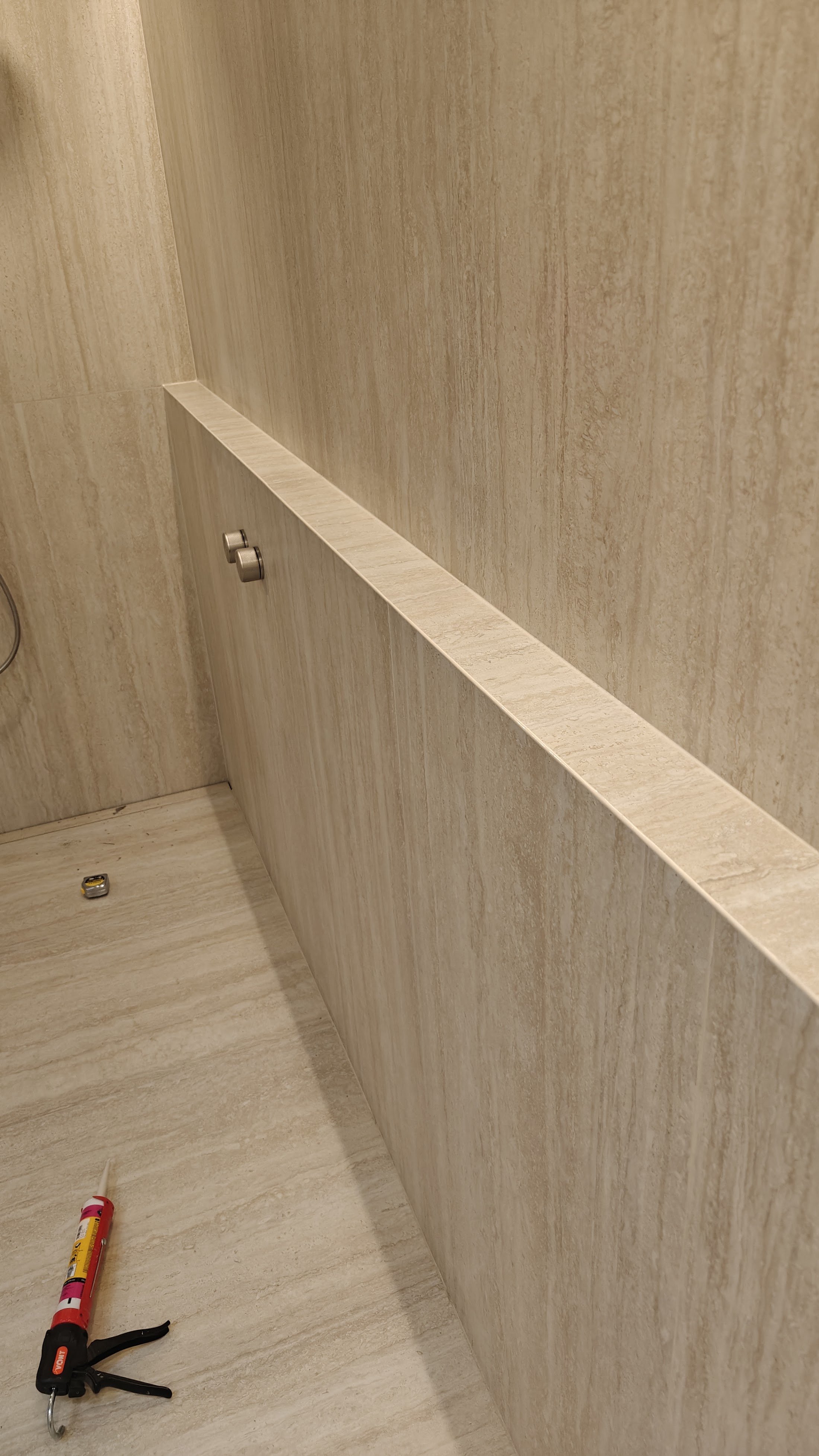 Bathroom with beige marble walls and floor, a white partition, a small cabinet with two metallic knobs, a caulking gun on the floor, and a small object near the corner.