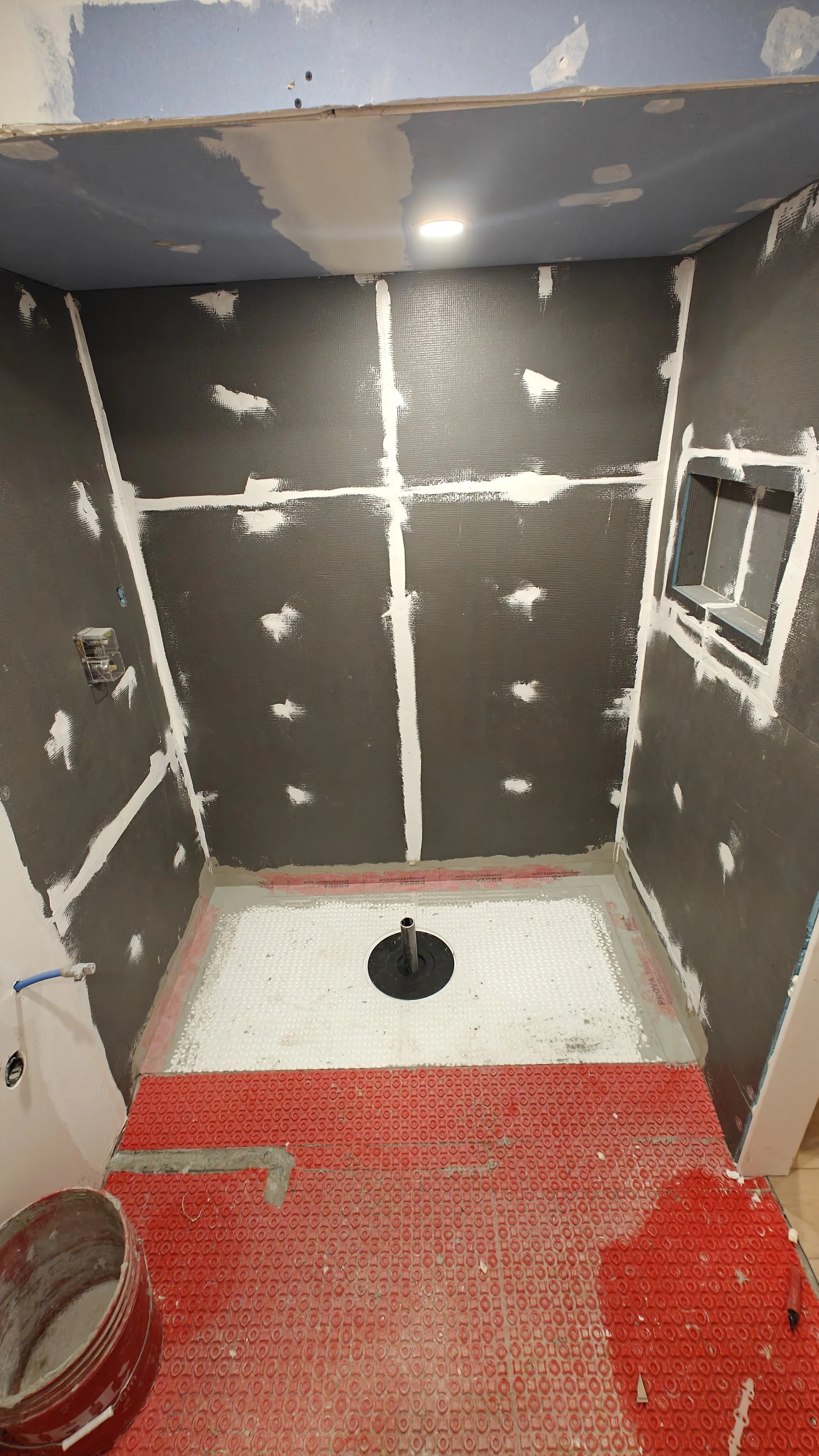 Under-construction bathroom with cement board walls, a circular plumbing fixture on the floor, and protective red floor covering. The ceiling has a recessed light, and there are small window openings on the walls.