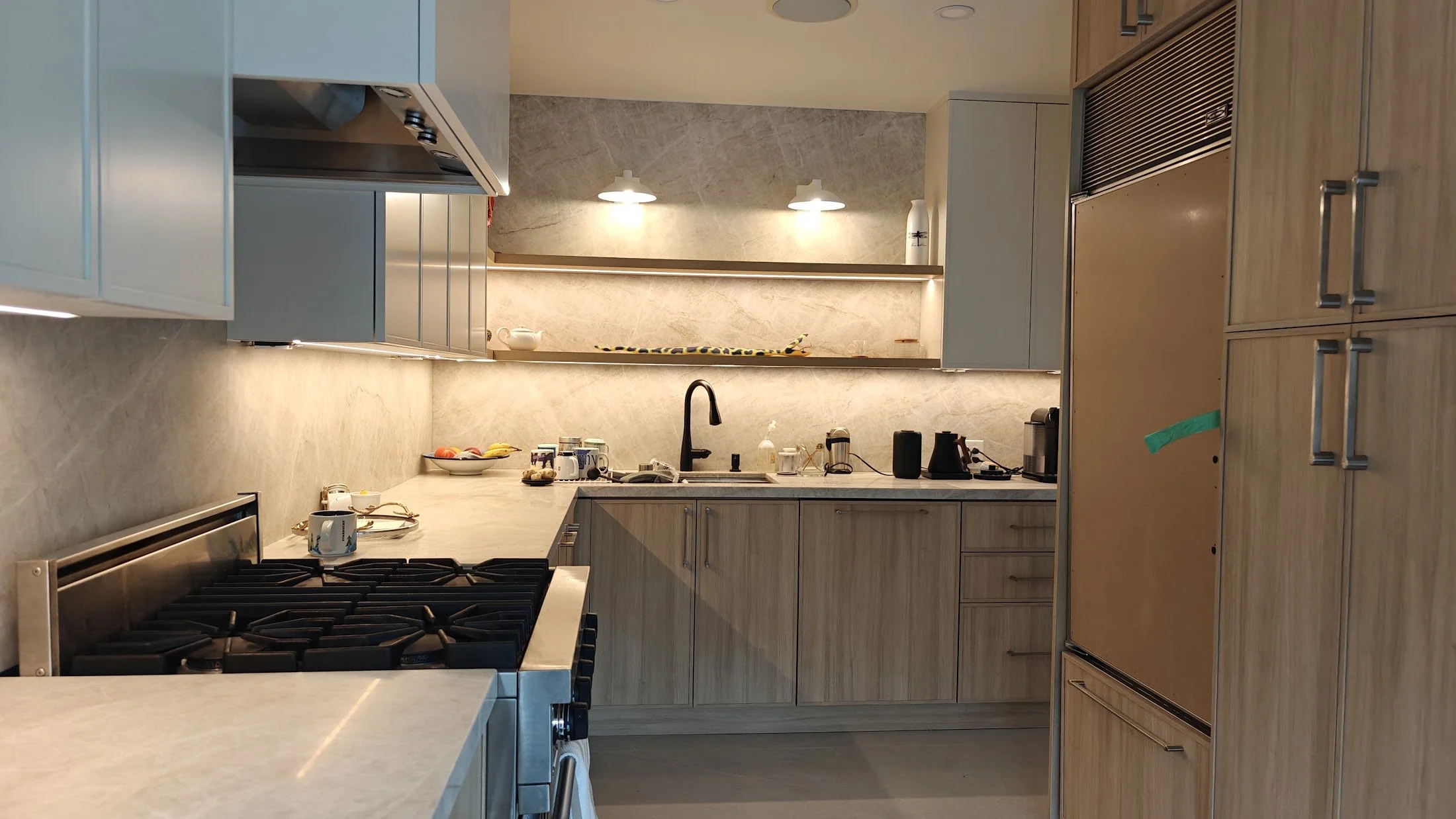 Modern kitchen with light wood cabinets, marble backsplash, and black appliances, cluttered with mugs, fruit, and small appliances.