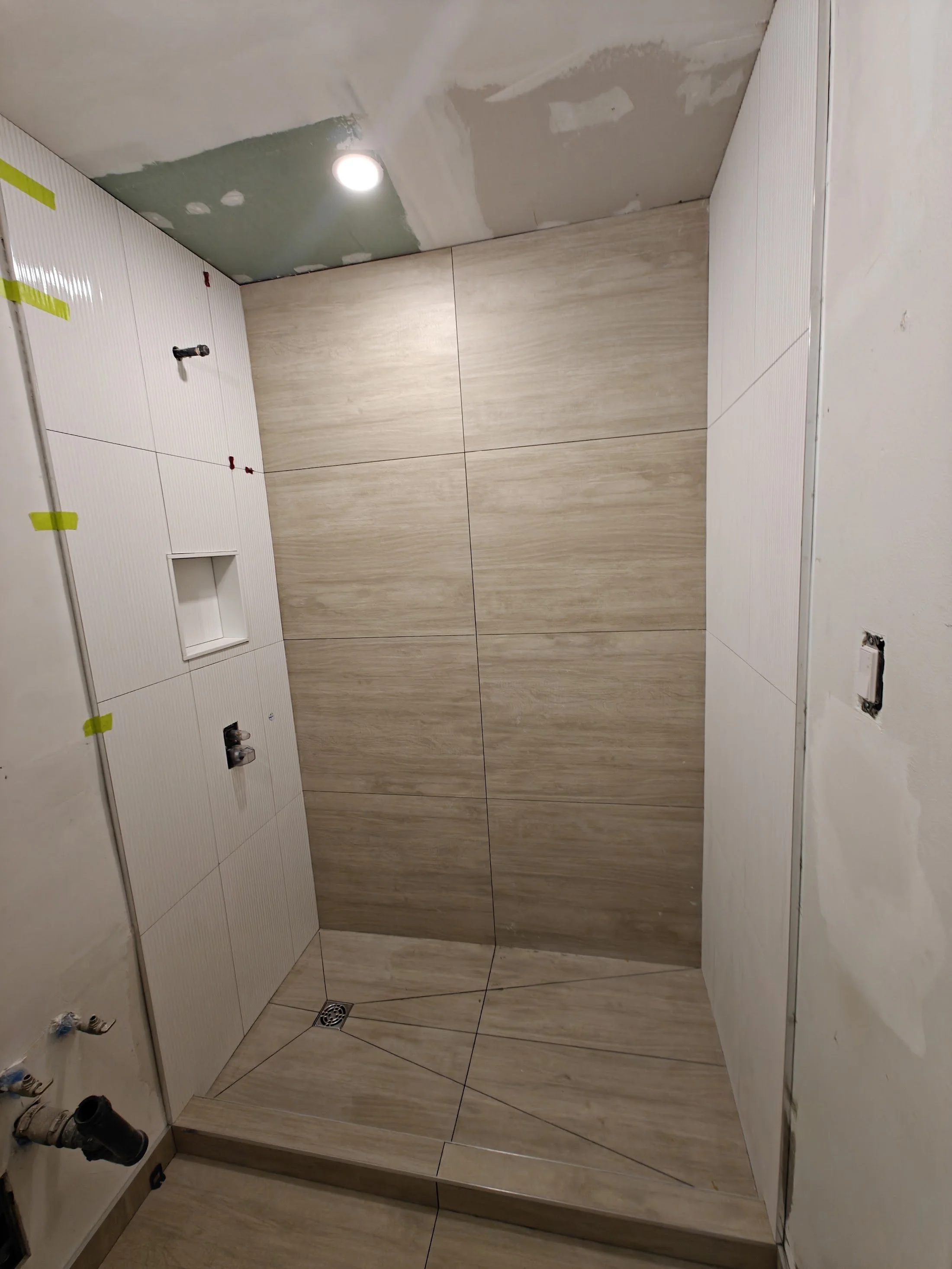 A bathroom under construction with beige wall tiles, white wall panels, a ceiling light, and unfinished plumbing fixtures.