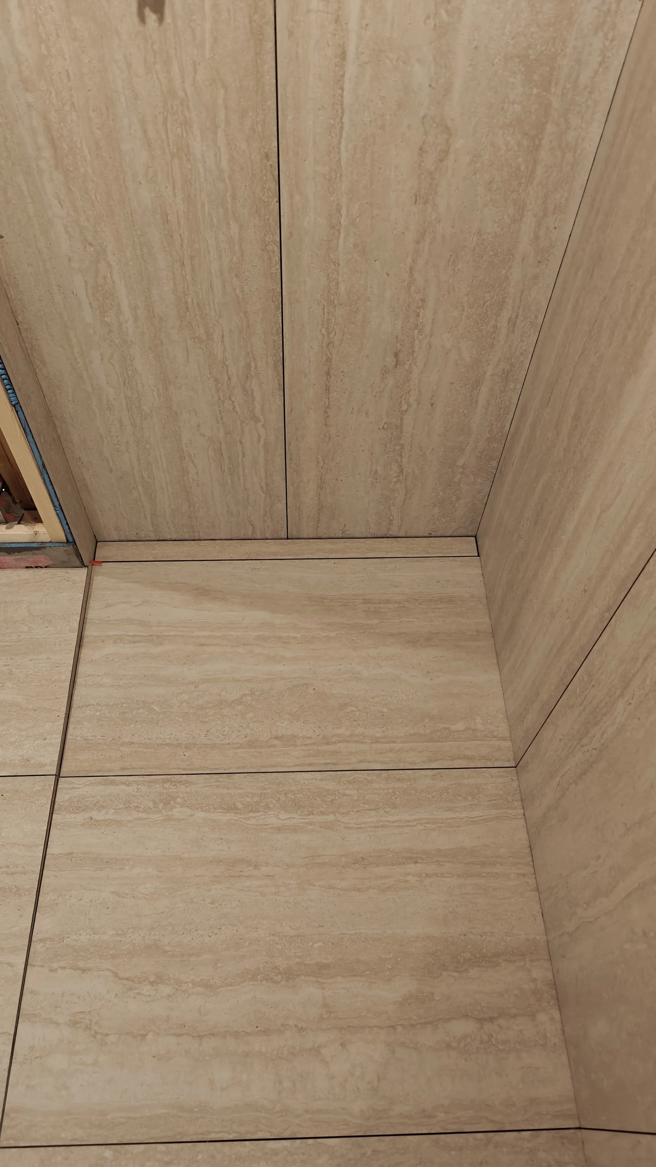 Corner of a room with beige marble tiles on the floor and walls, uninstalled baseboards, and a partially visible doorway.
